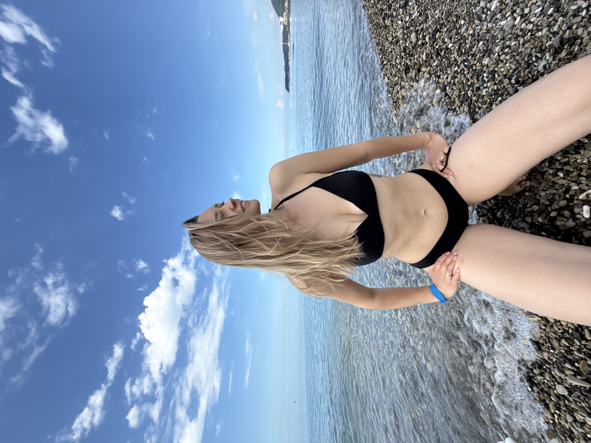 A blonde woman in a black bikini kneels on a pebbly beach in Nebug, Russia, with the Black Sea stretching out before her under a bright blue sky dotted with white clouds. The woman is facing right, looking out towards the horizon. The water gently laps at the shore, with small waves creating foam around her knees. In the distance, mountains and buildings are visible along the coastline.