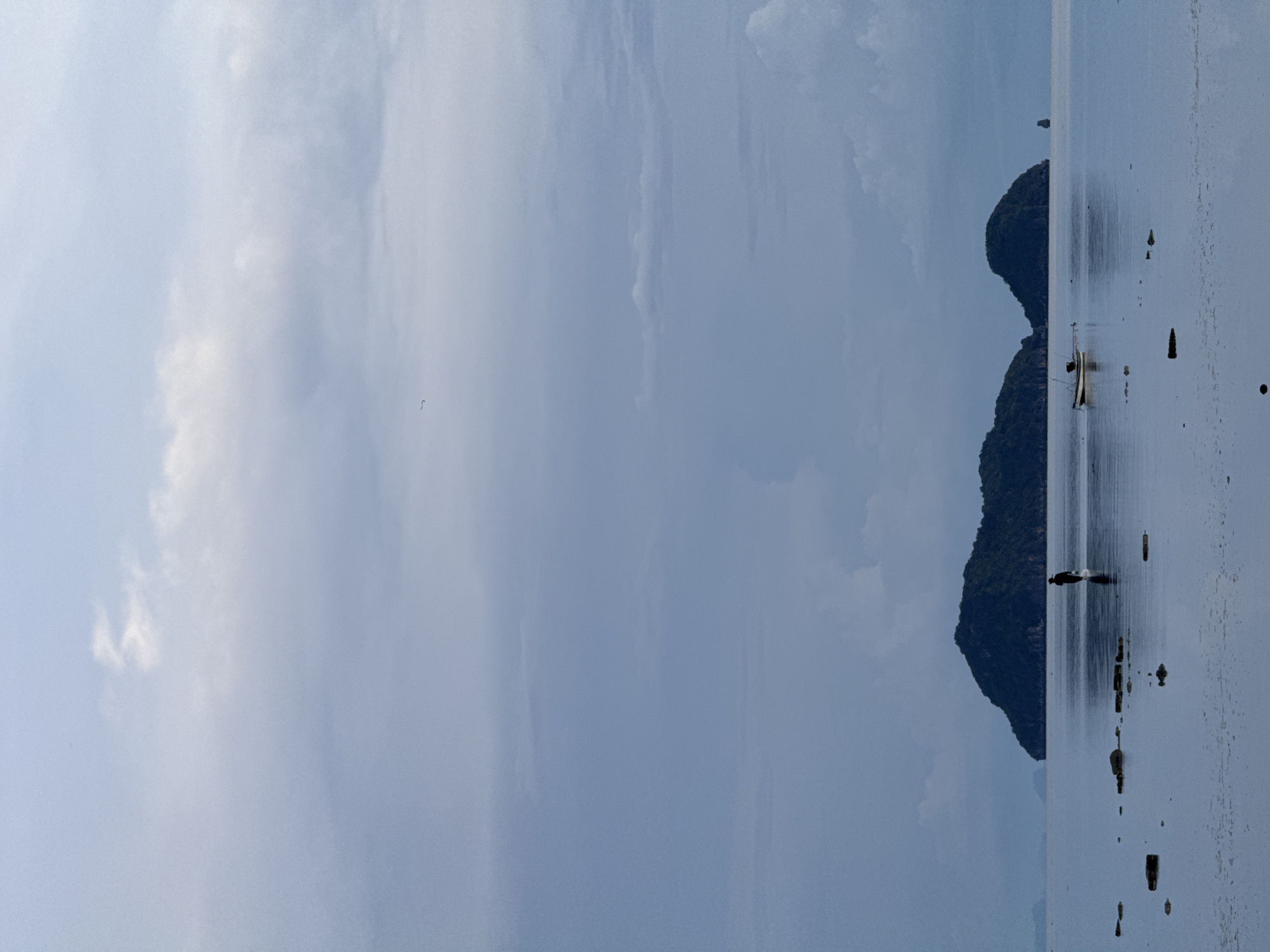 The image depicts a serene coastal scene with a solitary figure walking in shallow water near two small, verdant islands. The sky is overcast with soft, diffused light, contributing to a tranquil atmosphere. In the foreground, the calm water reflects the sky and the distant landmasses. Several dark, irregular shapes, likely rocks or debris, are scattered across the water's surface. A small, utilitarian boat is moored in the mid-ground, near the islands. The figure, silhouetted against the water, appears to be a lone individual engaged in an activity such as foraging or wading. The overall impression is one of peaceful solitude and natural beauty. This scene is consistent with the coastal landscapes found in ตำบลเกาะพะงัน, Thailand, known for its islands, beaches, and clear waters.