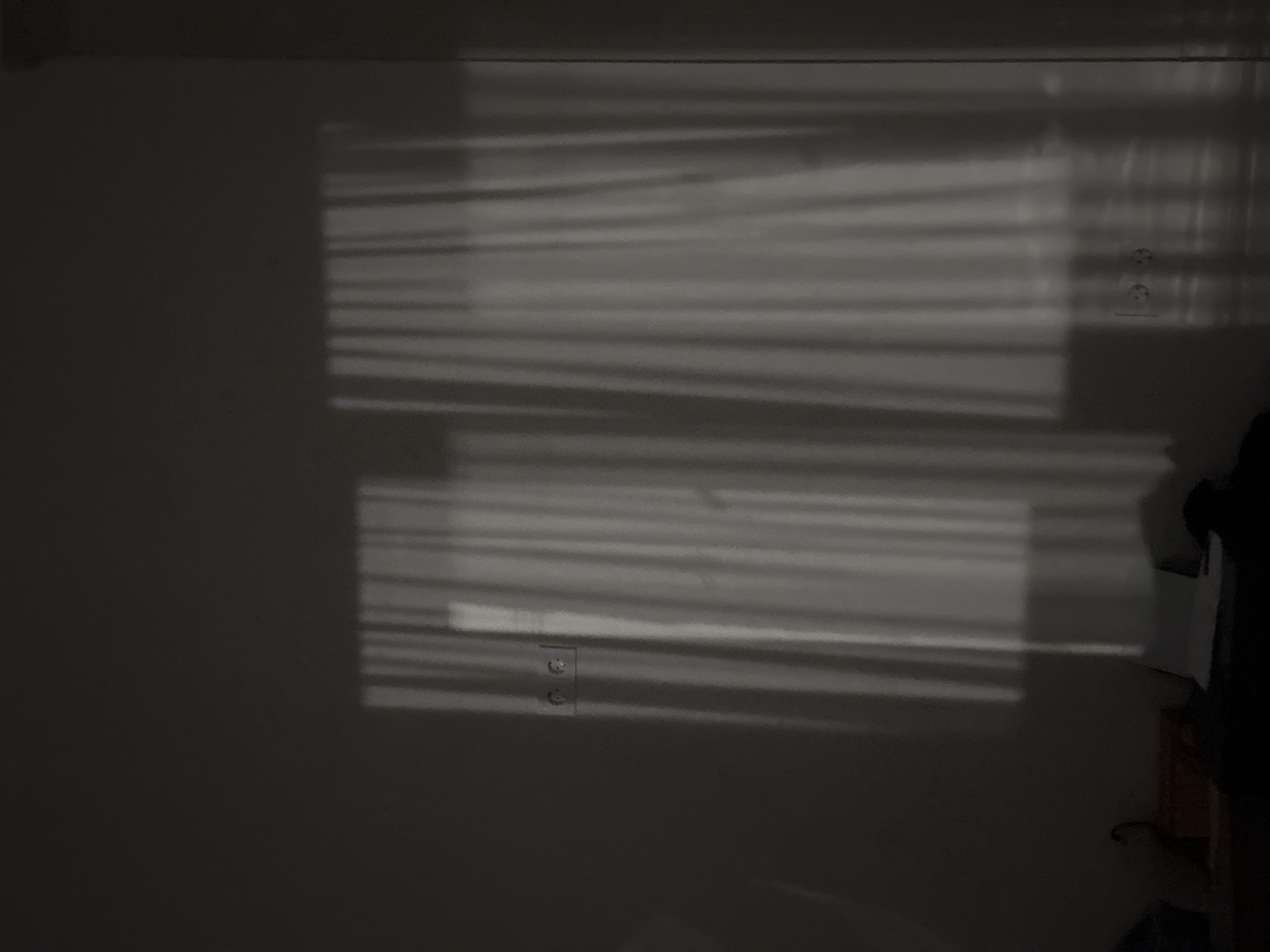 The image shows a wall with shadows cast by Venetian blinds. Two electrical outlets are visible on the wall, one in the lower left and one near the top right. Part of a dark object, potentially clothing, and a wooden structure are seen in the lower right corner. The location in the city is unknown.