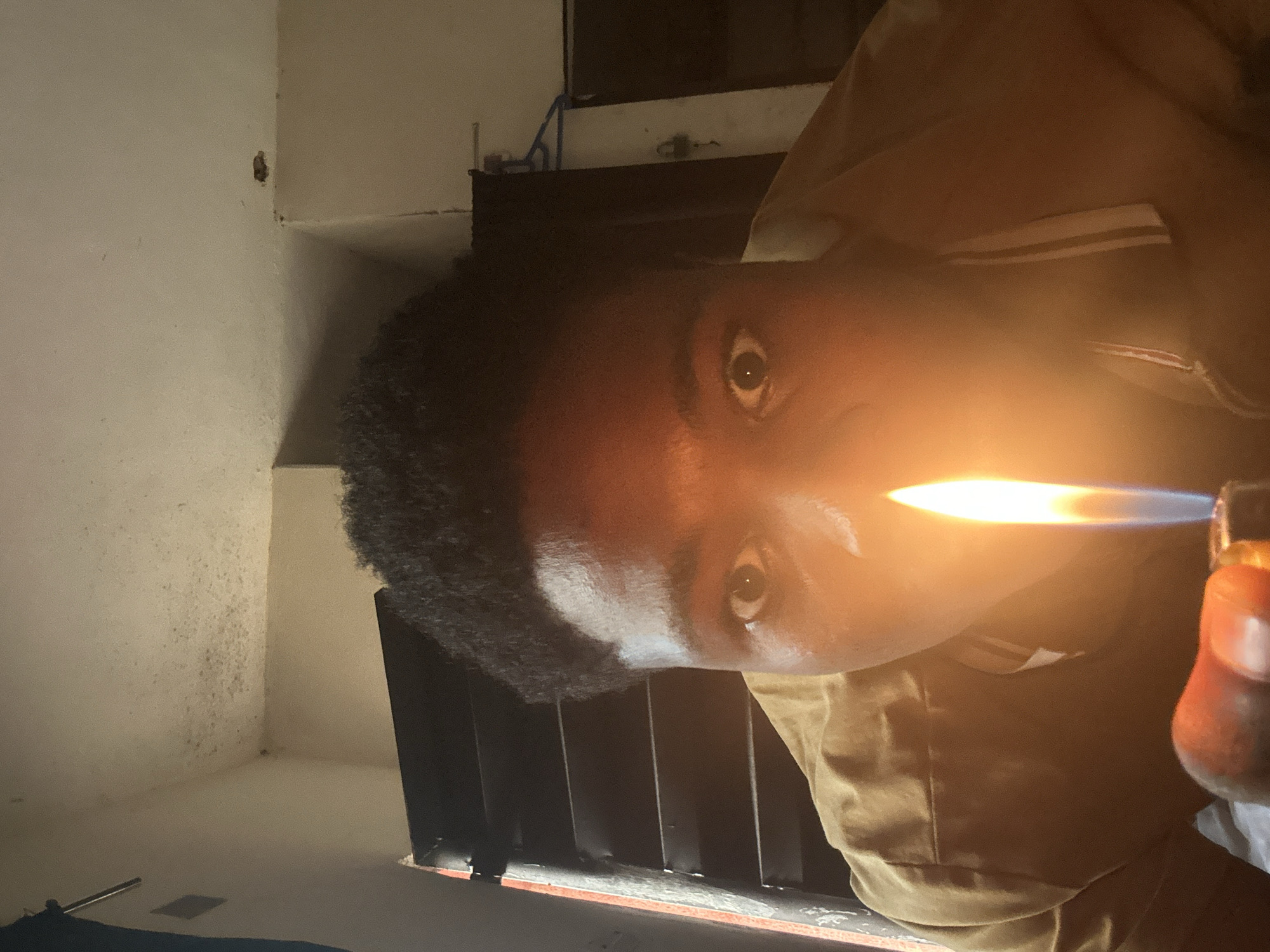 A close-up, low-angle shot shows a young Black person holding a lit lighter in front of their face. The flame illuminates their wide eyes and the front of their brown shirt. The background is a dimly lit room with horizontal blinds on the left and a light-colored wall on the right, possibly indicating a residential setting. There is some discoloration on the ceiling and walls. The image was captured indoors in Ihiagwa, Nigeria.