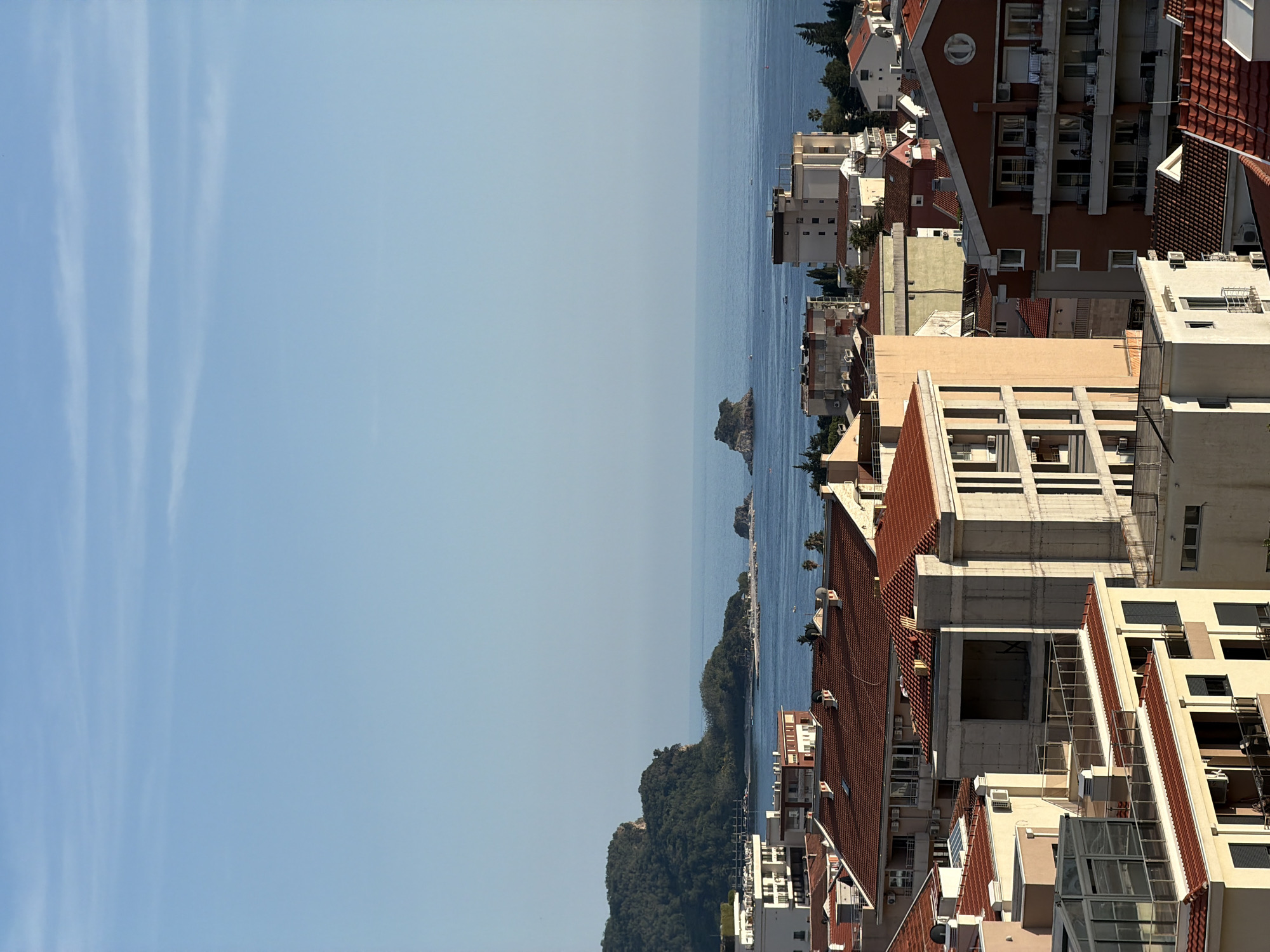 The image shows a view from above, likely from a high building, overlooking Budva, Montenegro. The foreground is dominated by the rooftops of buildings, mainly red-tiled, with white and beige structures interspersed. In the middle ground, a body of water stretches horizontally across the frame, with a small island or rocky outcrop visible. Beyond the water, the horizon meets a clear, light blue sky with faint streaks of clouds. Lush green vegetation covers the land along the water's edge on the right side of the frame. No people are visible.