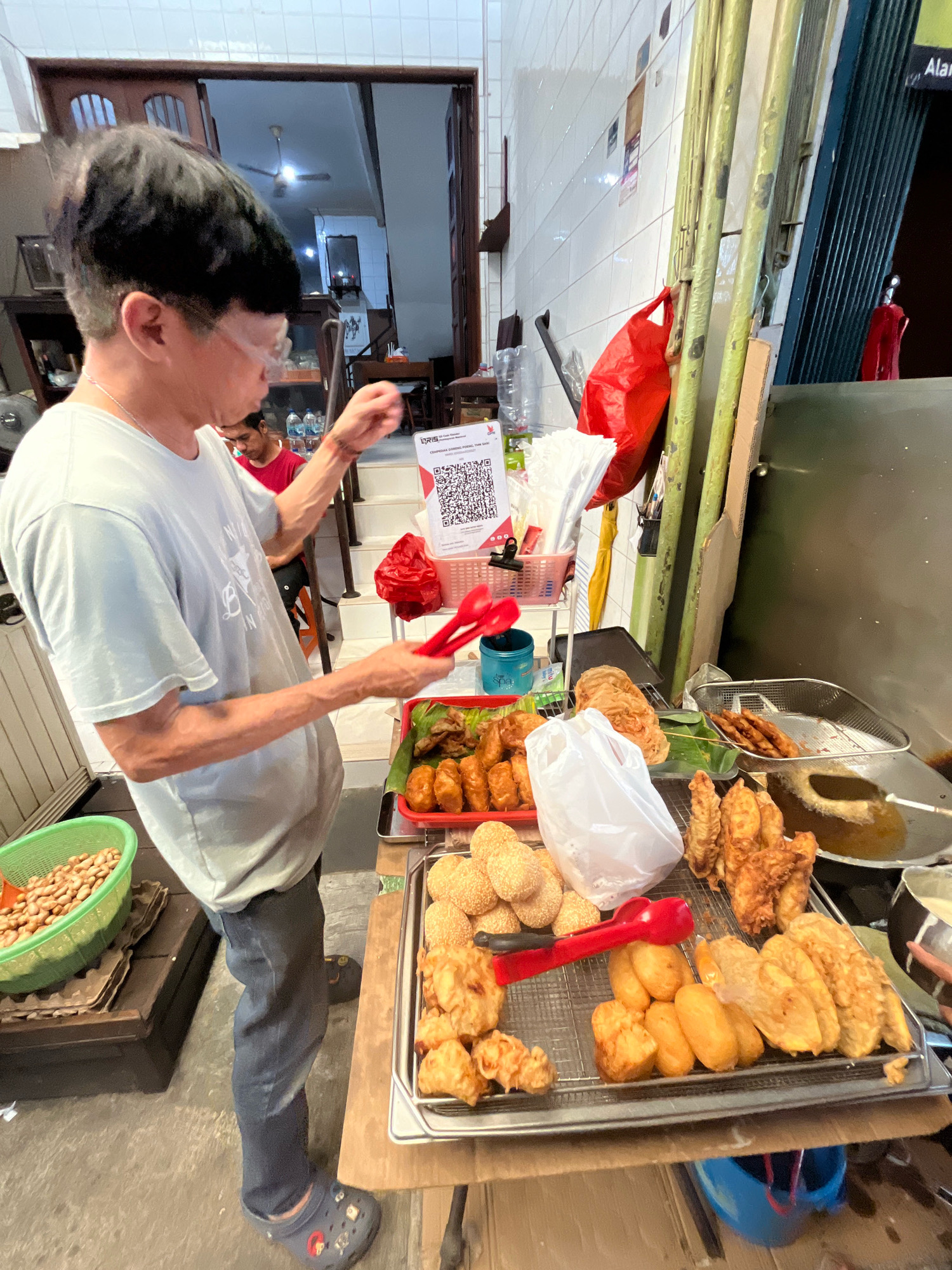 In an outdoor stall with a tiled wall and a metallic shutter, a man wearing a light blue t-shirt and dark pants stands behind a display of fried snacks. He is holding two red tongs in his right hand. The stall features a variety of fried items, including what appear to be bananas, pastries, and coated snacks, arranged on metal trays and a rack. To the left of the snacks, a green plastic basket filled with peanuts is visible. In the background, a staircase leads into a dimly lit interior. A QR code is displayed on a white stand, suggesting a payment option. The ground is paved with a rough concrete surface.