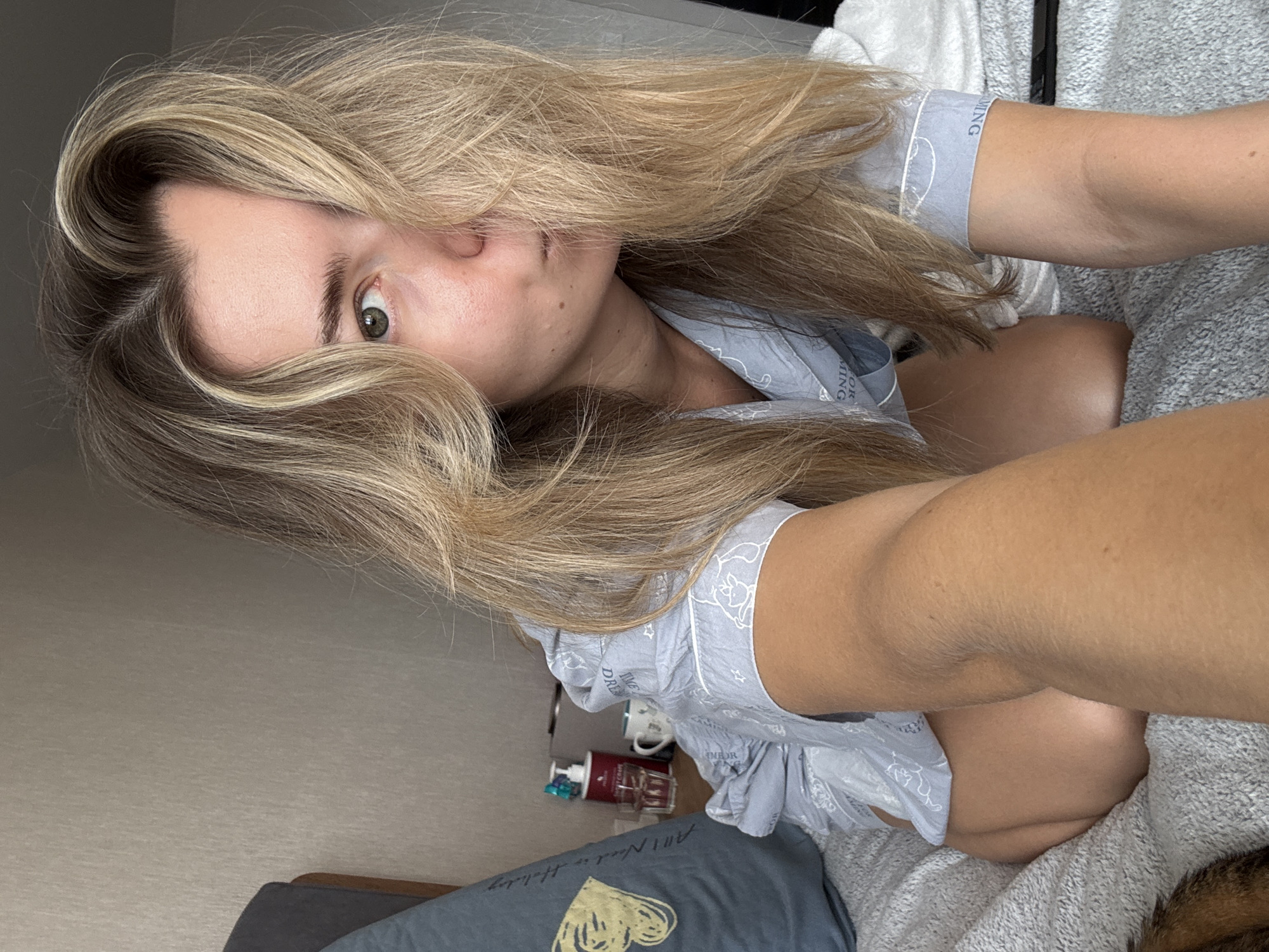 A young woman with blonde hair and green eyes is kneeling on a bed, looking towards the camera. She is wearing a light blue pajama set with a white pattern. Her hair is partially covering her face. To her left, a blue pillow with a yellow design is visible. On a nightstand behind her, there are a bottle of lotion, a glass of water, and a mug. The room appears to be a bedroom, with a textured wall in the background.