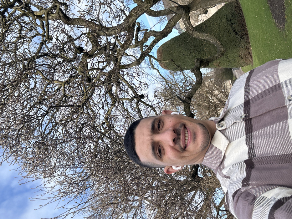 A male individual in his late 20s to early 30s is depicted taking a selfie in the Parc de l'Orangerie, Strasbourg, France. He occupies the lower-left foreground, smiling directly at the camera. He has short dark hair, a light stubble beard, and is wearing a white wireless earbud in each ear. His attire includes a long-sleeved flannel shirt with a plaid pattern in shades of beige, white, and brown. The background is dominated by large deciduous trees with bare, gnarled branches, indicative of late winter or early spring. To the right, a tall, precisely trimmed, dark green topiary, shaped like a tapered cone, stands on a vibrant green lawn. Distant classical architectural elements are faintly visible through the trees in the far background under a blue sky with scattered white clouds.