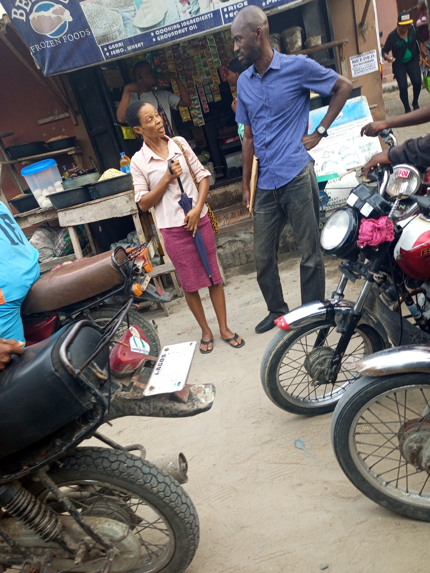 This media file captures a vibrant street scene at a market stall in Satellite Town, Nigeria, during daytime. In the foreground, two motorcycles are parked, with the one on the left showing a brown seat and red body, and its license plate clearly displaying "LAGOS". Another motorcycle on the right features a red and white tank, a chrome fender, and a headlight with a visible "FOR" sticker.
Two main individuals are featured in the midground. A woman wears a light pink collared shirt, a maroon skirt, and flip-flops, holding a dark umbrella. Her expression suggests she is speaking or reacting to something off-camera, with her mouth slightly open. Beside her, a bald man in a blue short-sleeved collared shirt, dark jeans, and dark shoes stands with his right hand on his hip, holding a light-colored paper or folder, looking left.
Behind them, a market stall or shop is visible, covered by a blue banner advertising "BE FROZEN FOODS". The banner also lists items like "GARRI", "SEMO, WHEAT", "COOKING INGREDIENTS", and "GROUNDNUT OIL". Below the banner, shelves hold various goods, including a blue container with eggs, black trays filled with what appear to be grains, and numerous small packaged items hanging. A person is partially visible behind the counter, wearing an apron. On the right side of the stall, a sign is partially legible, stating "KITCHEN ASSISTANT NEEDED IN A RESTAURANT". Another person with a blue t-shirt is visible on the left motorcycle, and a person in a yellow cap walks away in the background. The ground is unpaved, appearing sandy or dusty.