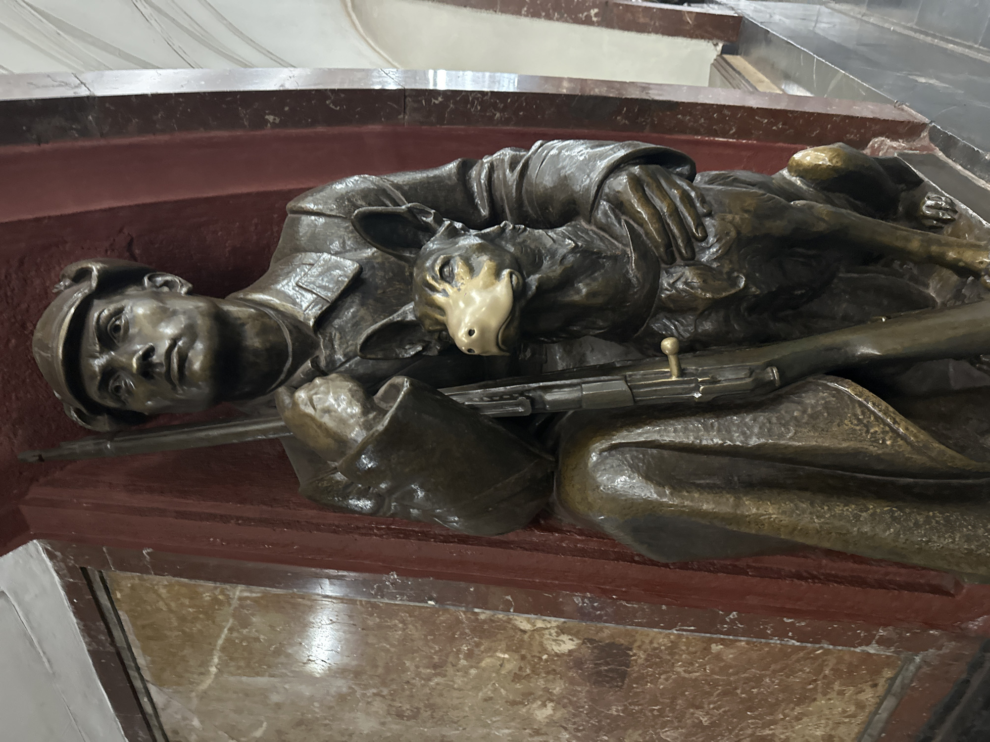 This is a bronze sculpture located in the metro station Mayakovskaya in Moscow, Russia. The sculpture depicts a man in a cap and coat, holding a rifle. Beside him, a dog sits with its head turned to the left. The sculpture is situated against a backdrop of marbled walls. The man's face is solemn, and his gaze is directed upwards and to the left. The dog's mouth is slightly open, and it appears alert. The overall color palette of the sculpture is metallic bronze with lighter highlights, and the background features earthy tones and marbled textures. The lighting suggests an indoor environment, characteristic of a metro station.