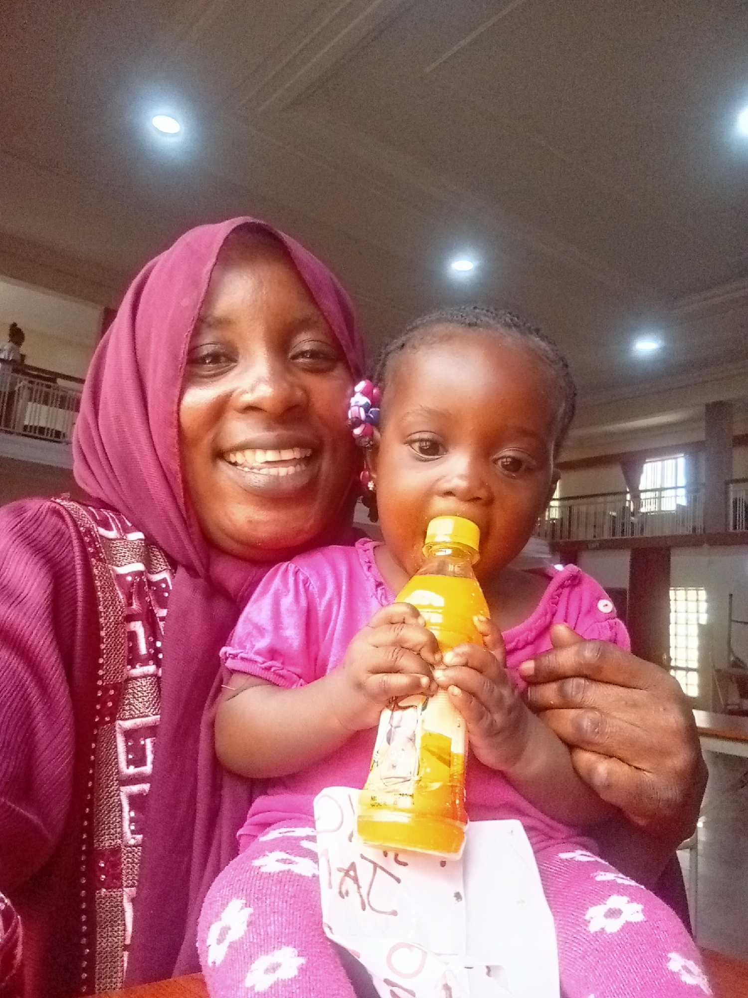 A woman wearing a maroon hijab and a maroon patterned garment is smiling at the camera. She is holding a young child on her lap who is drinking from a bottle of yellow liquid. The child is wearing a pink outfit and pink patterned socks, and there is a white object, possibly a piece of paper, underneath her. The background shows a building interior with a balcony and what appears to be a window with bars. Based on the architecture and cultural context, it is likely that the image was taken in Akwanga, Nigeria.