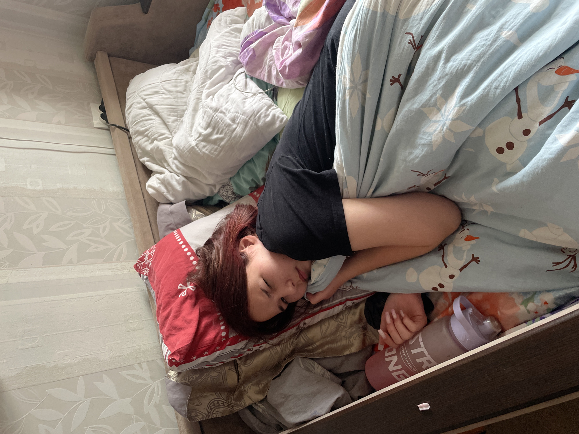 A young woman is depicted sleeping or resting in a bed, viewed from a slightly elevated, diagonal perspective. She is lying on her left side, facing away from the camera, with her head resting on a red pillow patterned with white snowflakes or stars. Her reddish-brown hair is visible around her head. She is wearing a black t-shirt and is largely covered by a light blue duvet featuring white Olaf characters and snowflakes from the movie Frozen. Her arms are tucked close to her body under the duvet, though one hand with long, light-colored artificial nails is visible near a water bottle.
The bed is filled with various layers of bedding, including a white quilted comforter, a purple patterned sheet, and other blankets and pillows, suggesting a cozy, lived-in space. A light pink plastic water bottle with a light purple cap is placed beside the woman within the bed frame, which appears to be a light brown wood or upholstered material. Visible text on the water bottle includes "STRON" and "OVE THE WISDOM".
The room's walls are adorned with light-colored wallpaper that has a subtle, repeating pattern of leaves or vines. An electrical outlet with a black cable plugged in is visible on the wall behind the head of the bed. The overall scene conveys a sense of peaceful rest in an indoor setting, likely a bedroom. The lighting suggests daytime, but there are no specific cues regarding the time of day, weather, or exact location. The woman appears calm and asleep.