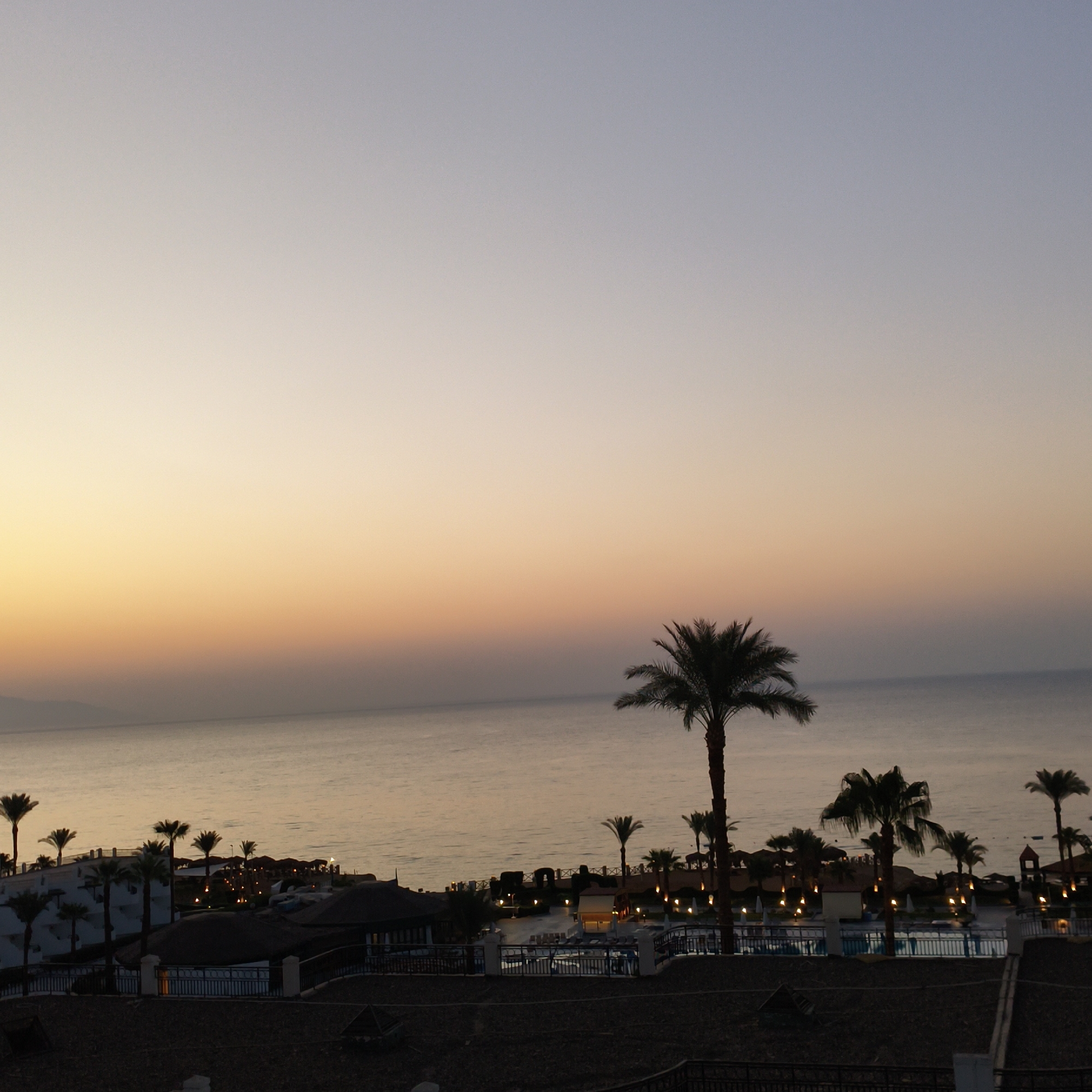 The image depicts a resort scene at dusk or dawn, with a gradient sky transitioning from pale yellow and orange near the horizon to a soft blue overhead. The calm sea stretches out to the horizon, with a distant landmass visible on the left. In the foreground and mid-ground, there are several palm trees silhouetted against the sky and the developing twilight. A resort complex with multiple buildings, swimming pools, and outdoor lighting is visible along the coastline. The buildings appear to be multi-story structures with white walls and some thatched roofs. The lighting from the resort indicates that it is operational. The overall atmosphere is tranquil, suggesting a peaceful evening or morning at a tropical or coastal destination.