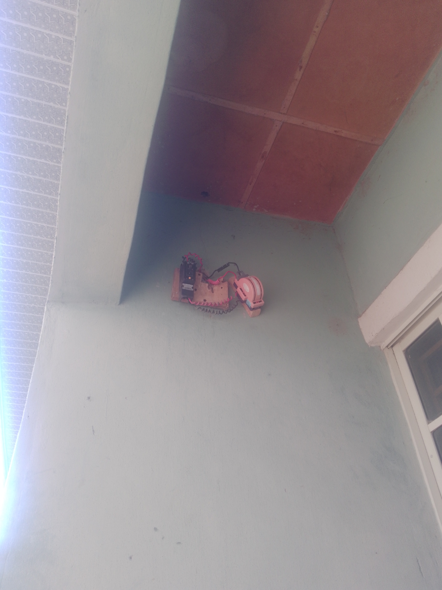The image captures an outdoor scene, likely a wall in Jauro Yinu, Nigeria. Mounted on a light blue wall is a electrical component, possibly a meter or a junction box. It is mounted on a wooden board and features several wires and a red lever or switch. To the left of the electrical component, a decorative pattern is visible on what appears to be a tiled or textured surface. Above the wall, a reddish-brown ceiling with visible support beams can be seen. To the right, part of a white-framed window is visible. The overall impression is one of an external electrical fixture on a building.