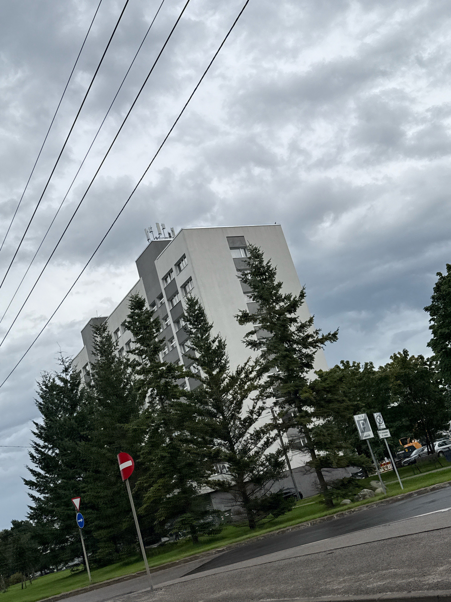 The image depicts a view of a modern apartment building with a light grey and white facade, featuring multiple windows and a row of telecommunication antennas on its rooftop. Several tall, dark green evergreen trees stand in front of the building, partially obscuring it. In the foreground, a wet asphalt road curves to the right, with a grassy area and sidewalks alongside. Several traffic signs are visible, including a circular red sign with a horizontal white bar, and a triangular white sign with a blue arrow. The sky is overcast with heavy, dark grey clouds. Power lines are visible in the upper left quadrant of the image, running diagonally across the frame. In the background, other trees and glimpses of other buildings and vehicles can be seen, including what appears to be a yellow construction vehicle. The perspective is from a low angle, looking upwards towards the building and the sky. The overall impression is of a typical urban residential area on a cloudy day, likely in Tallinn, Estonia, given the presence of distinctive traffic signs common to the region.