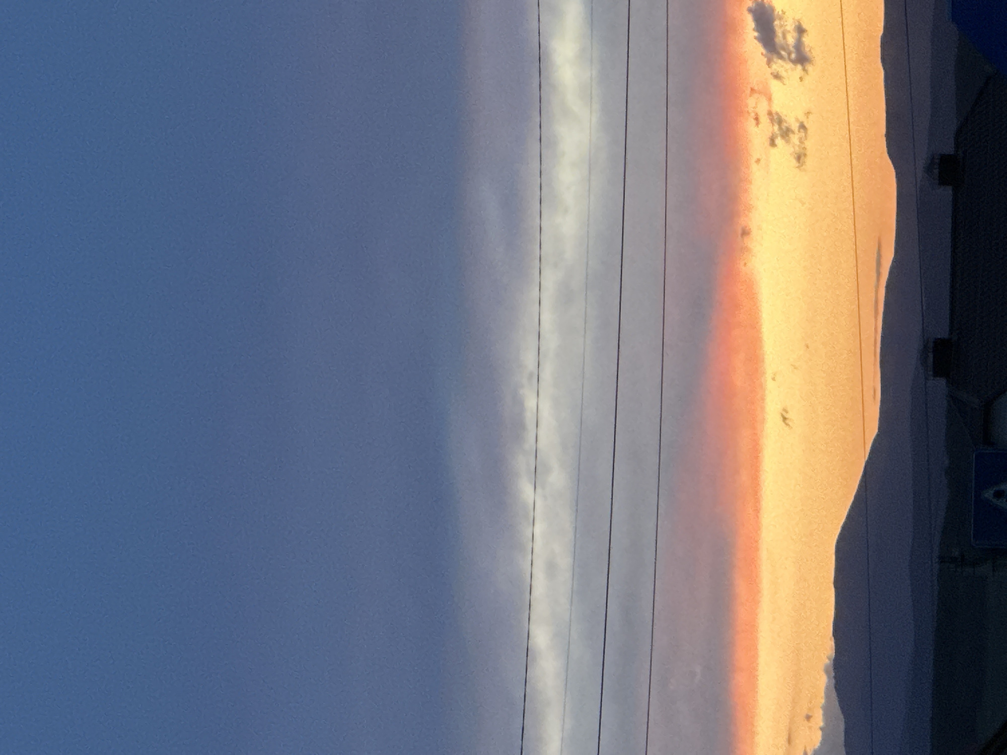 The image depicts a late afternoon or sunset sky with visible power lines. A building is partially visible on the right edge. The sky is transitioning from a deep blue on the left to a vibrant orange and red on the right, indicative of sunset colors. There are some clouds partially lit in the orange area of the sky. A sign, possibly a road sign, is attached to the building.
Based on the query, the location is identified as Uchaly, Russia, but there is no specific information to confirm this.