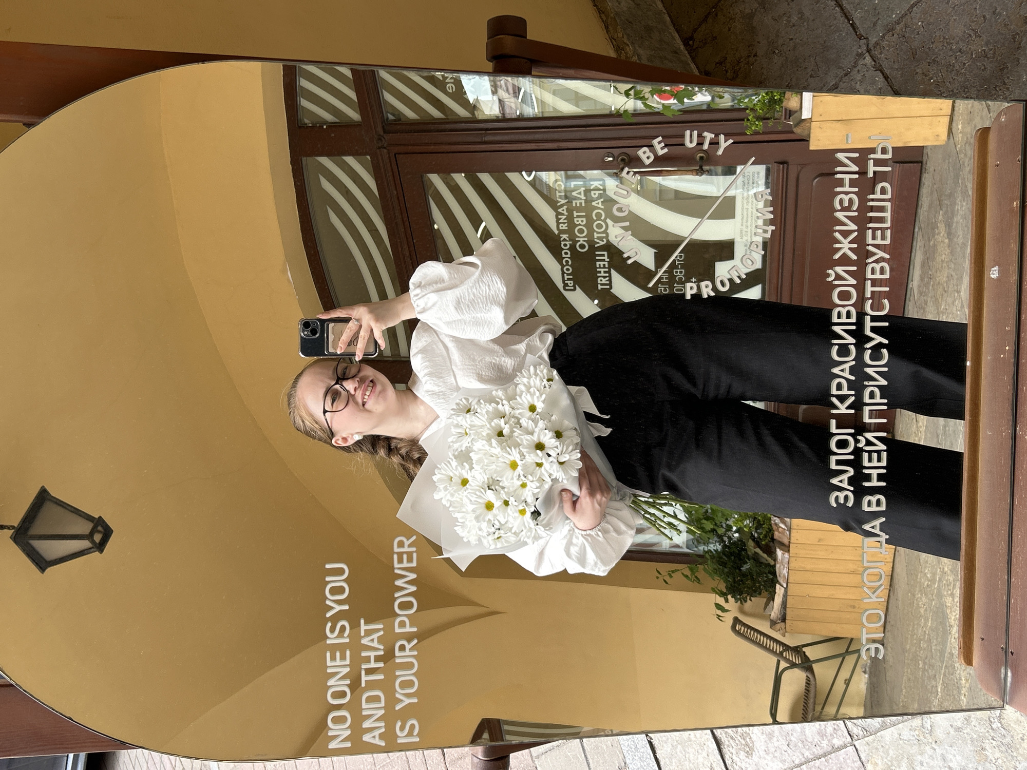 A young woman wearing a white blouse, black pants, and glasses is taking a selfie in a large, oval-shaped mirror. She is holding a bouquet of white daisies in her left hand. The mirror reflects a building with a striped awning and a sign that reads "PROPORCIYA". The location is the city of Saint Petersburg, Russia. The mirror also has text written in both English and Russian. The English text reads, "NO ONE IS YOU AND THAT IS YOUR POWER". The Russian text translates to "The key to a beautiful life is when you are present in it".