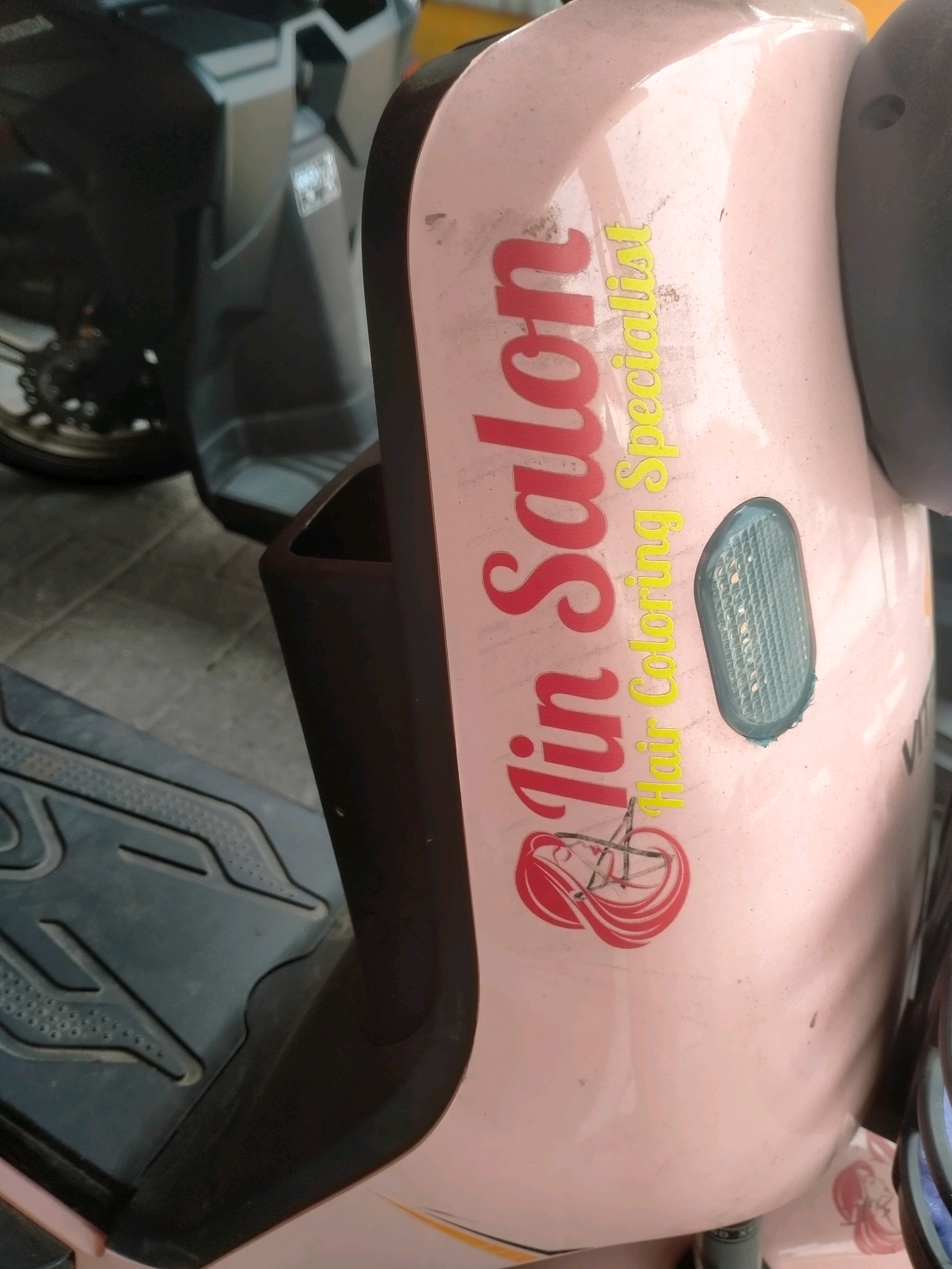 A close-up shot captures a pink scooter in Kasihan, Indonesia. The scooter's side panel features a red and yellow sticker with the words "Jin Salon" and "Hair Coloring Specialist" in stylized fonts. A graphic of a woman's head with flowing hair and scissors is also part of the sticker. In the background, the dark gray body of another scooter is partially visible, and a paved surface with scattered debris can be seen on the ground. The lighting suggests it is daytime, with some dirt and dust present on the scooter's surface.