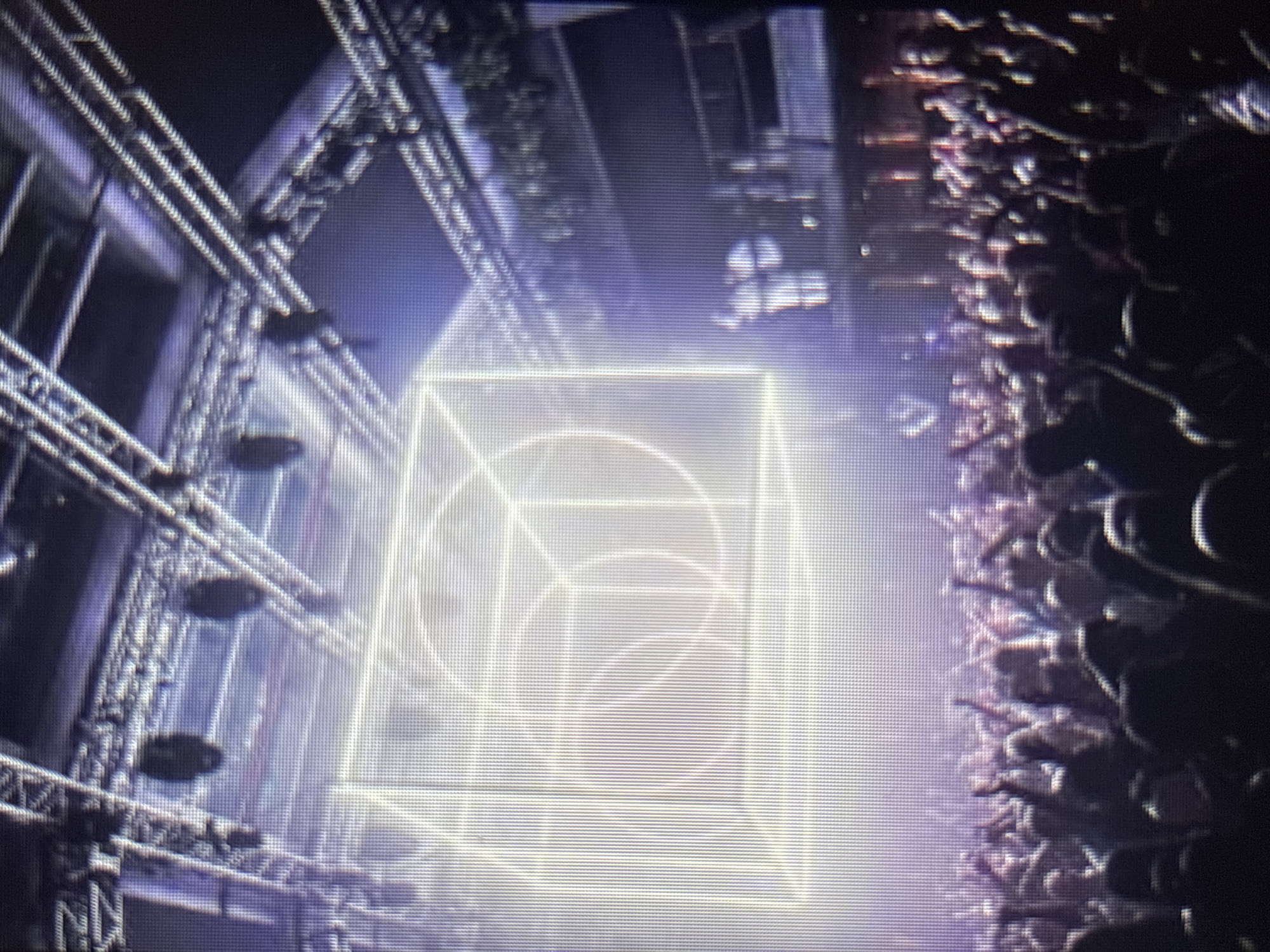 The image depicts a lively indoor concert or event venue. A large, illuminated cube-like structure, outlined with bright lights and featuring geometric patterns including concentric circles and lines, is centrally positioned and serves as a focal point. Above, a complex industrial-style lighting rig with numerous spotlights and trusses is visible. Below the illuminated structure, a dense crowd of concert-goers is gathered, with many people raising their hands in the air, suggesting an energetic and engaging atmosphere. The lighting is predominantly purple and white, creating a dramatic and atmospheric effect. The overall impression is that of a modern entertainment space hosting a vibrant event.
