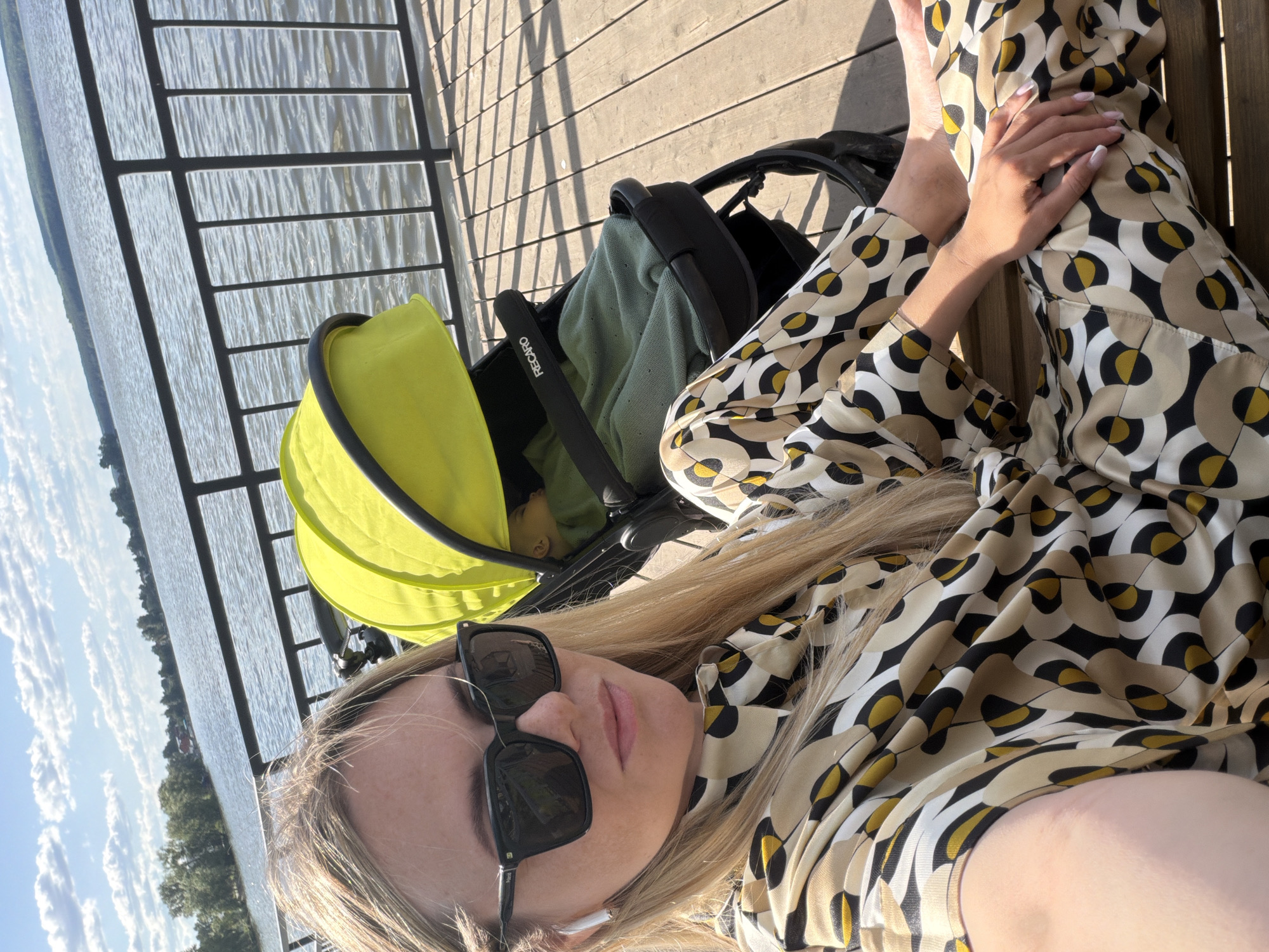 A selfie-style image depicts a blonde female and an infant in a stroller situated on a wooden pier overlooking a substantial body of water in Votkinsk, Russia. The female, in the foreground, wears dark square-framed sunglasses, one white wireless earbud in her left ear, and a matching patterned shirt and trousers featuring a geometric design in black, beige, white, and yellow. Her right hand rests on her lap. To her right, a black Recaro baby stroller with a bright lime green canopy shades an infant, whose upper face is partially visible beneath the canopy, covered by a mint green knitted blanket. The wooden pier consists of parallel planks and is bordered by a dark metal railing separating it from the calm, rippling water. The background reveals a distant shoreline with green trees and scattered buildings, under a blue sky adorned with white cumulus clouds. Direct sunlight illuminates the scene, casting distinct shadows on the pier's wooden surface.