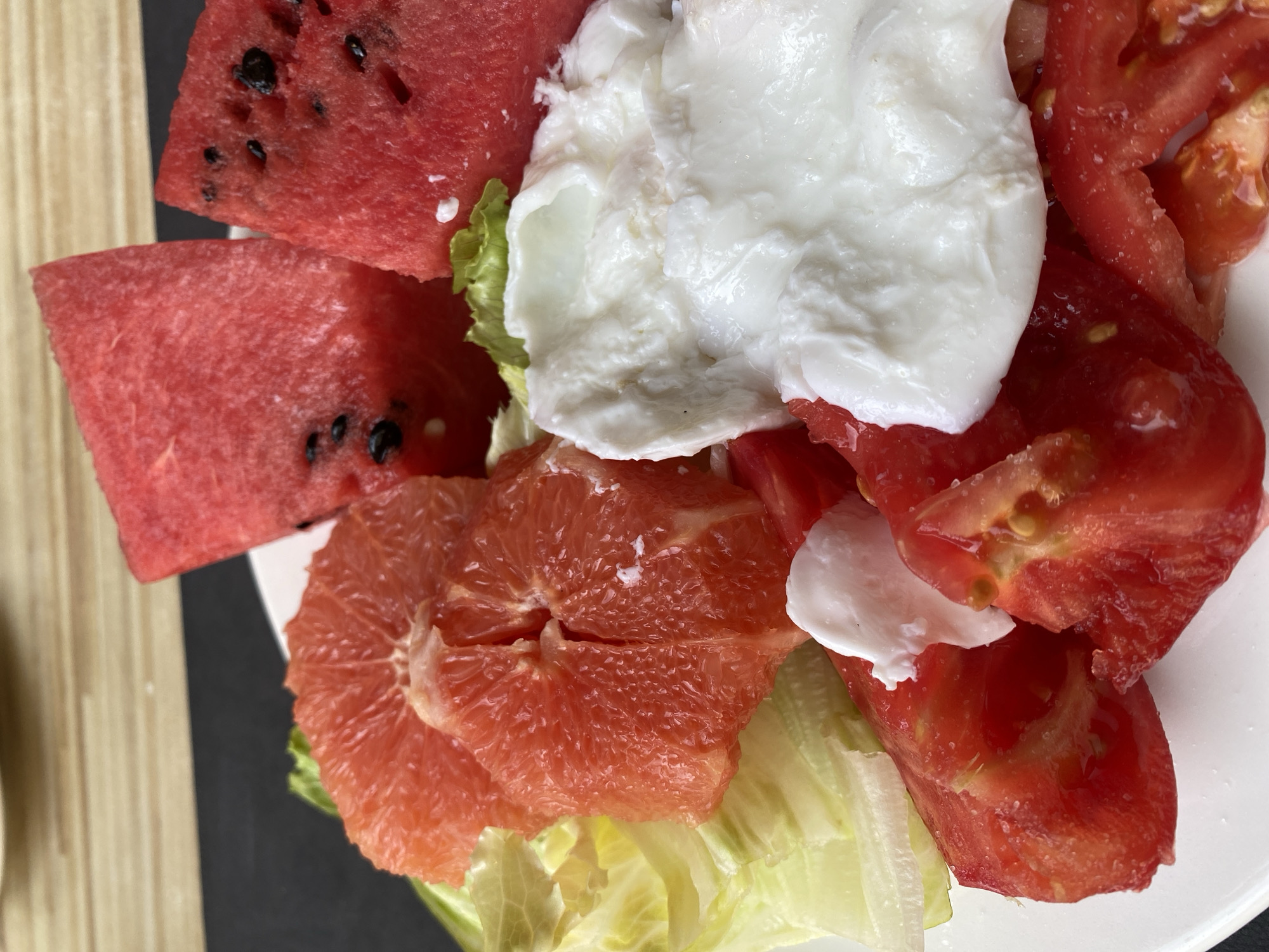 A close-up, overhead shot features a white plate piled high with a colorful salad. The salad is composed of cubed watermelon, segmented grapefruit, chopped tomatoes, fresh lettuce, and a dollop of fresh mozzarella cheese. The background is blurred, showing a dark surface and a hint of a wooden table. There are no people visible in the image, and the focus is entirely on the food. The image appears to have been taken in Aksu, Turkey.