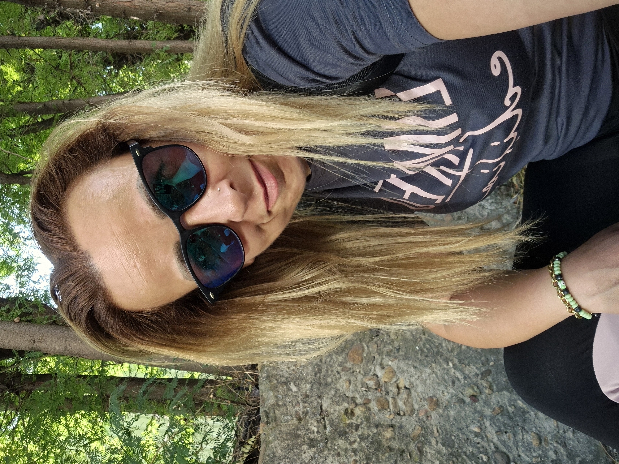 A woman with blonde hair and sunglasses is posing for a picture outdoors. She is wearing a dark t-shirt with pink lettering and black pants. Her left arm is resting on her leg, and she is wearing a bracelet on her wrist. The background consists of trees and a stone wall.
