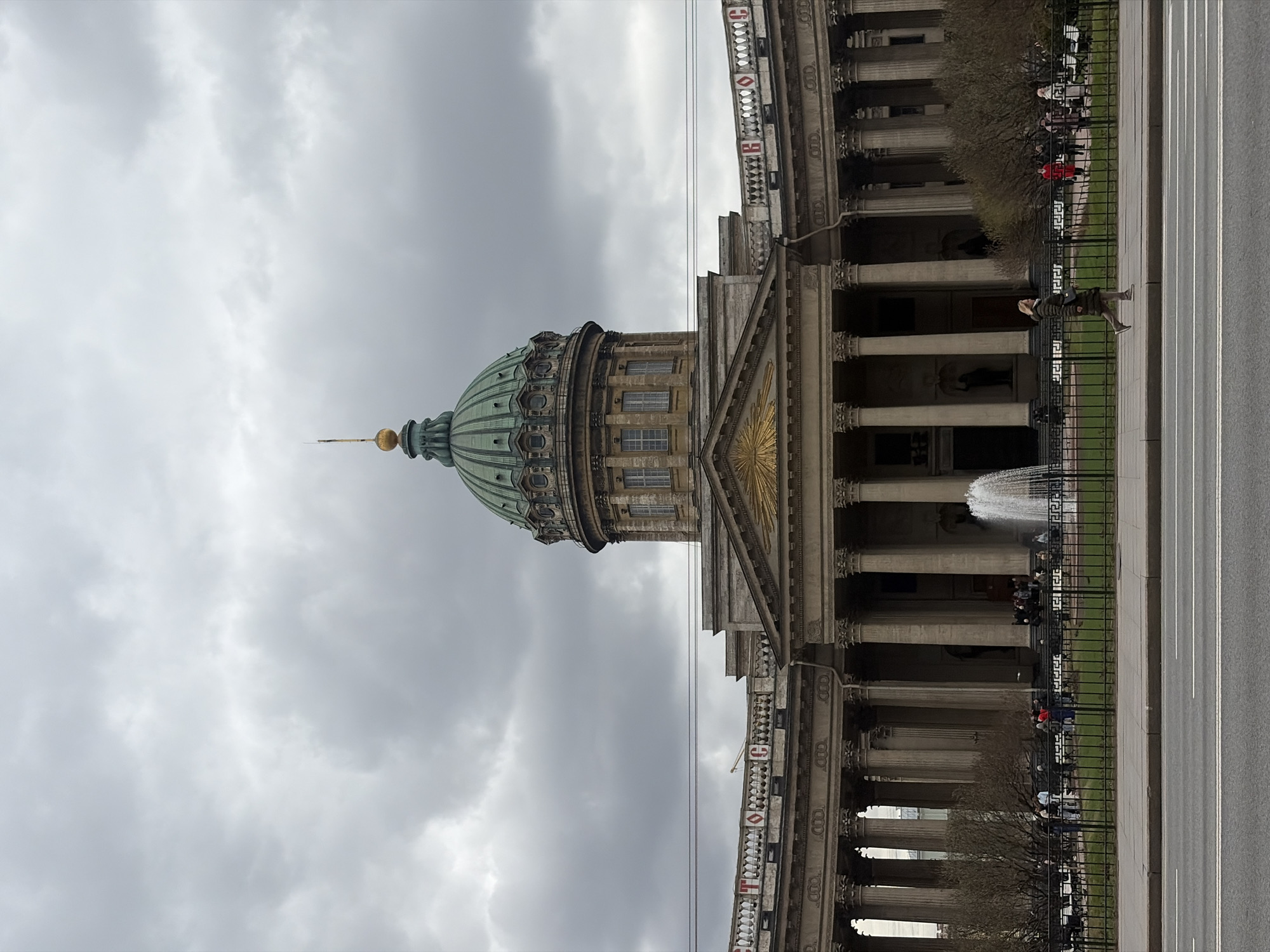 The image, when correctly oriented, captures the grand facade of the Kazan Cathedral in Saint Petersburg, Russia, under an overcast sky. The impressive Neoclassical architecture is the central subject, dominated by its large, fluted green dome, which is capped with a golden finial. Beneath the dome, a prominent colonnade with numerous classical columns supports a triangular pediment adorned with a golden sunburst relief. A decorative balustrade runs along the top of the building, featuring red and white elements with visible fragmented letters such as "C", "O", "T", "I", and "E".
In the middle ground, a black metal fence separates a narrow strip of green grass and some bare trees from a wide paved sidewalk. Further right, a multi-lane asphalt road with painted white lines extends across the frame. A fountain actively sprays water high into the air in front of the cathedral, near the fence line.
Several people are scattered throughout the scene: one individual in a light-colored coat and dark pants walks along the sidewalk, while others are seen sitting or standing further back, near the trees and cathedral entrance. The weather appears to be a cloudy daytime, with a uniformly grey sky suggesting no direct sunlight or bright conditions. The bare trees indicate either early spring or late autumn. The scene portrays a typical urban day around a significant architectural landmark, without any specific event taking place.