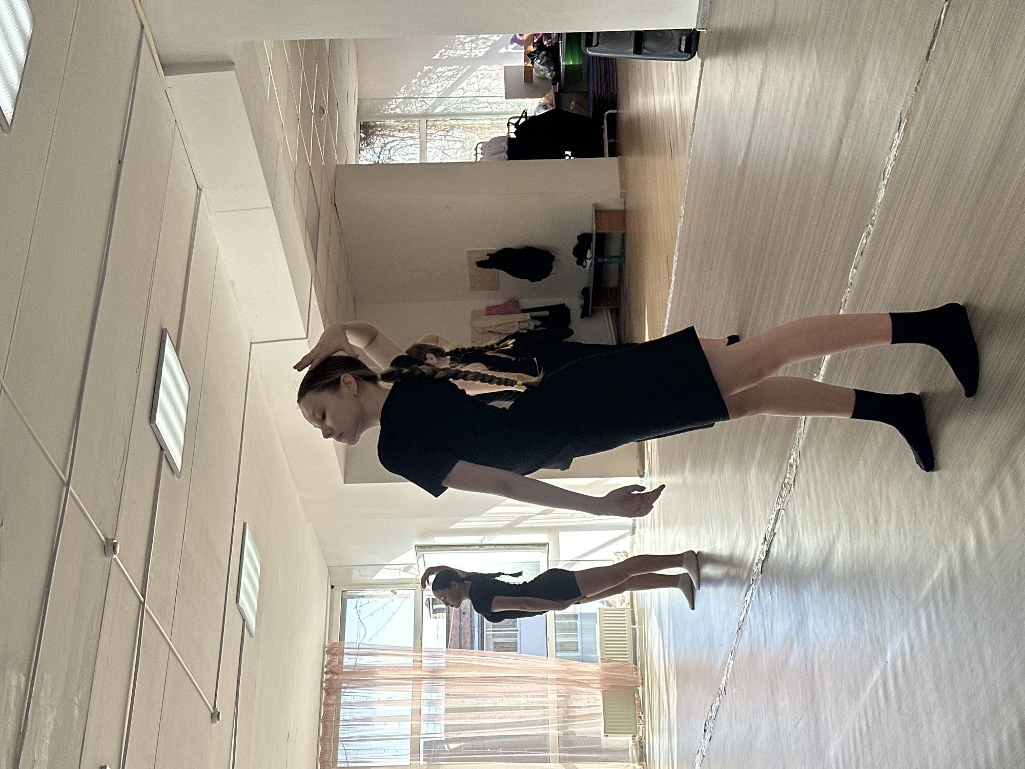 A young woman is in a dance studio in Votkinsk, Russia. She is wearing a black dress and socks, and her hair is in braids. She is striking a pose, with one arm raised above her head and the other extended in front of her. There is a reflection of her in the shiny floor. The room is spacious and well-lit, with several large windows. There are other items in the background, including clothing and bags. The scene appears to be a dance rehearsal or practice session.