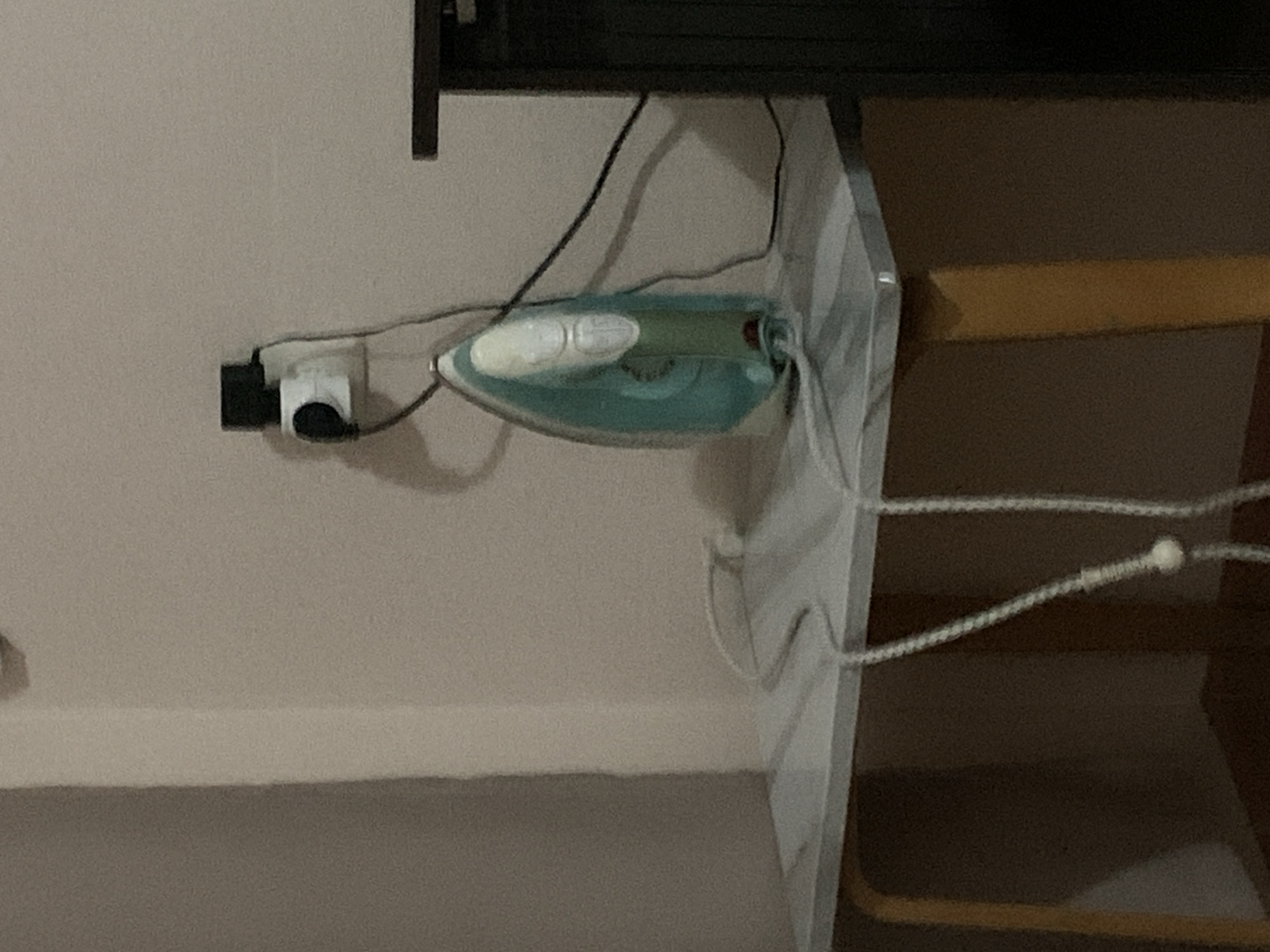 The image captures an indoor scene, likely a room in a home or hotel in Stavropol, Russia. The primary focus is an electric iron and its electrical connections.
A light blue and white electric steam iron is positioned face down on a flat, light-colored surface, possibly a shelf or a table. To the left of the iron, a white electrical wall socket is visible on a light-colored wall. A dark-colored plug, possibly an adapter or part of a larger power supply, is inserted into the outlet, with black cables extending from it towards the iron. The iron's braided power cord, which is light in color, extends towards the right of the frame, showing some slack.
Above the iron and cables, a dark, rectangular structure, possibly a television stand or another piece of furniture, spans across the upper portion of the image. To the far right, a section of furniture with a distinct yellowish-brown wooden element is partially visible, suggesting a chair or another table. A light-colored, possibly striped, piece of fabric is draped over the edge of the surface where the iron rests, near its handle.
No specific activity is taking place; the iron is merely resting, plugged into the wall. The dim lighting suggests it might be evening or that artificial light sources are being used. No people are present, and there is no visible text. The image's original orientation is rotated 90 degrees counter-clockwise, meaning the wall appears as the floor and the horizontal surface as vertical.