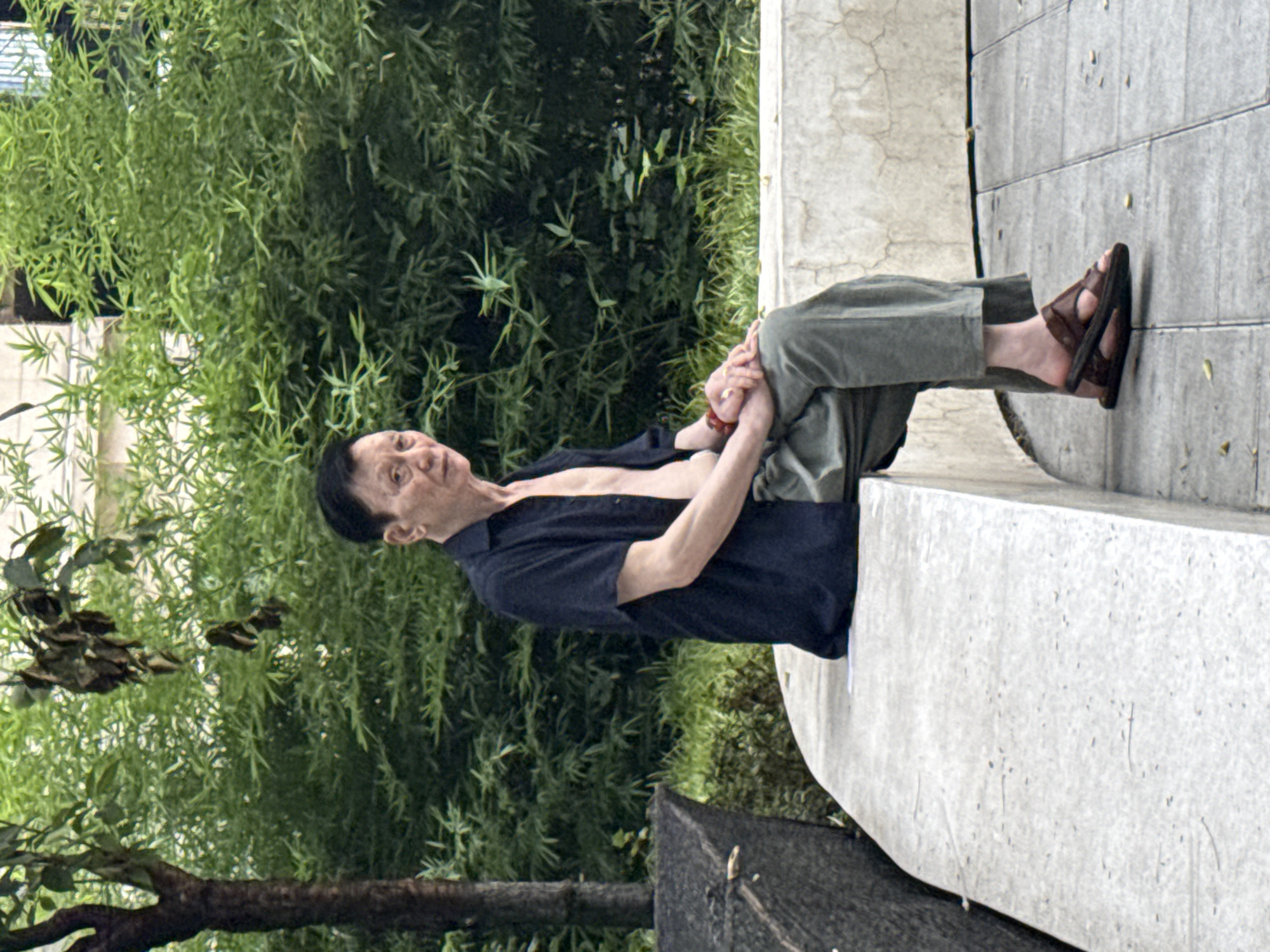 An elderly Asian man with short black hair and a wrinkled face sits on a curved, light-colored concrete bench in what appears to be a park or public space. He is wearing an unbuttoned dark blue short-sleeved shirt, revealing his chest, and loose-fitting green trousers. His right hand rests on his lap, while his left hand is clasped over his right. He wears brown leather sandals. Behind him, a dense wall of green bamboo foliage and a tree trunk are visible, suggesting an outdoor setting in a temperate climate. The bench is positioned on a tiled surface. The man is looking towards the right of the frame. The overall scene suggests a moment of rest or contemplation. The location is Yuzhong District, China.