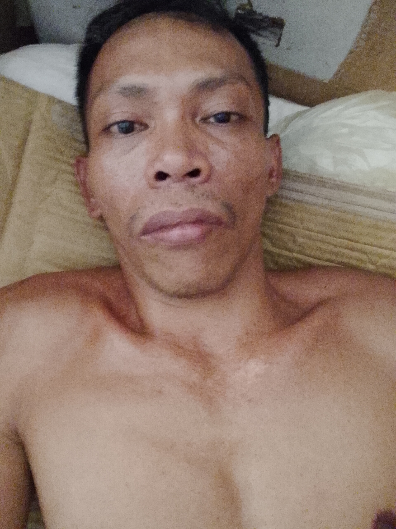 A shirtless man with short black hair is lying on a bed. He has dark skin, a mustache, and is looking directly at the camera. The background consists of a tan pillow and bedding. There is no indication of the location in Bukit Ketok, Indonesia.