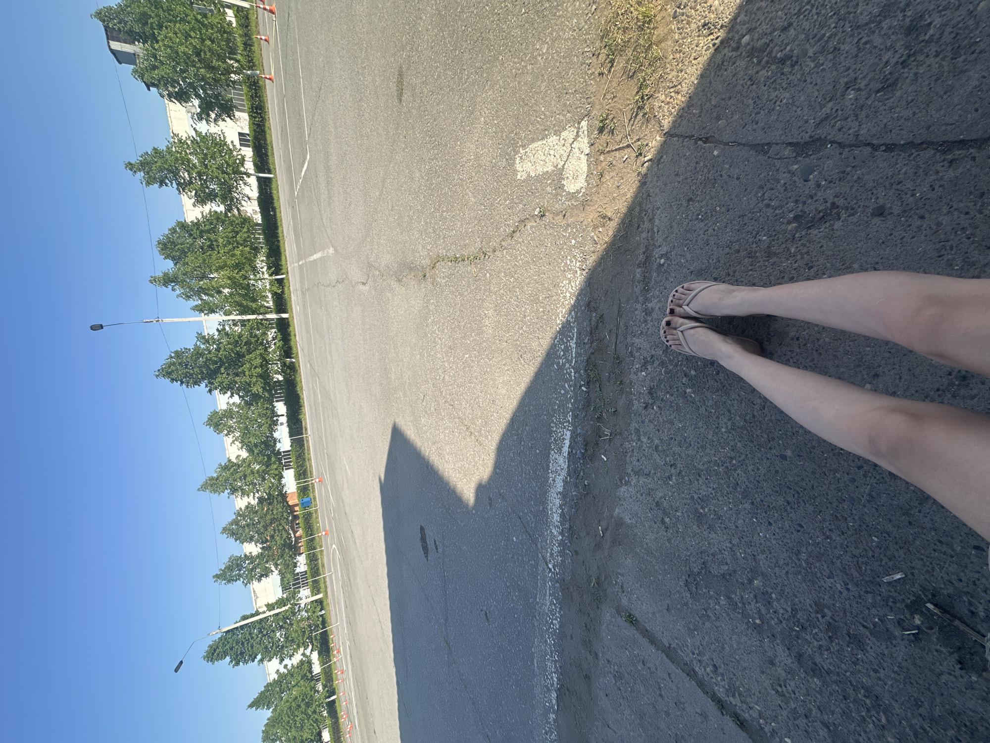 The image, taken in daylight, shows a partial view of an asphalt surface with painted white lines, typical of a parking lot or roadway. A person's legs and feet wearing flip-flops are visible in the lower right corner of the frame. The asphalt surface exhibits a clear demarcation between a sunlit area and a shadowed area, suggesting a strong, directional light source, likely the sun. Adjacent to the asphalt, there is a strip of ground with sparse vegetation and debris.
In the background, trees line the edge of the asphalt. The image appears to be taken in Chaikovsky, Russia, based on the style of buildings visible in the distance, the street lamps, and the general landscaping style which are congruent with architectural and infrastructure elements common in Russian urban environments.