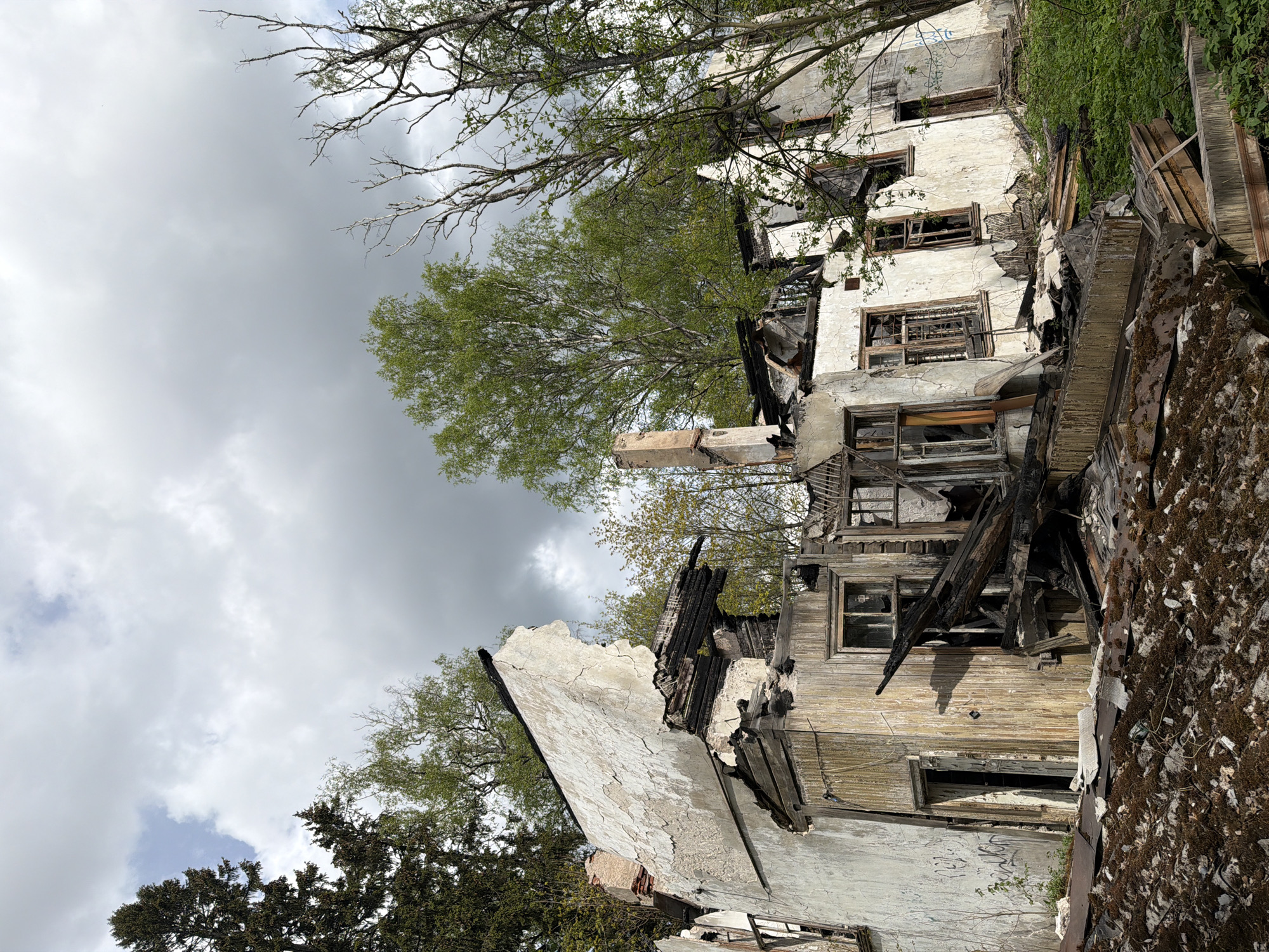The image shows a dilapidated building, possibly in a state of ruin due to fire damage and neglect. The structure is in Kolbino, Russia. Portions of the building are visibly charred with exposed wooden beams. The walls are crumbling and have graffiti. Windows are broken or missing. Surrounding vegetation includes trees and moss-covered debris. The sky is overcast.