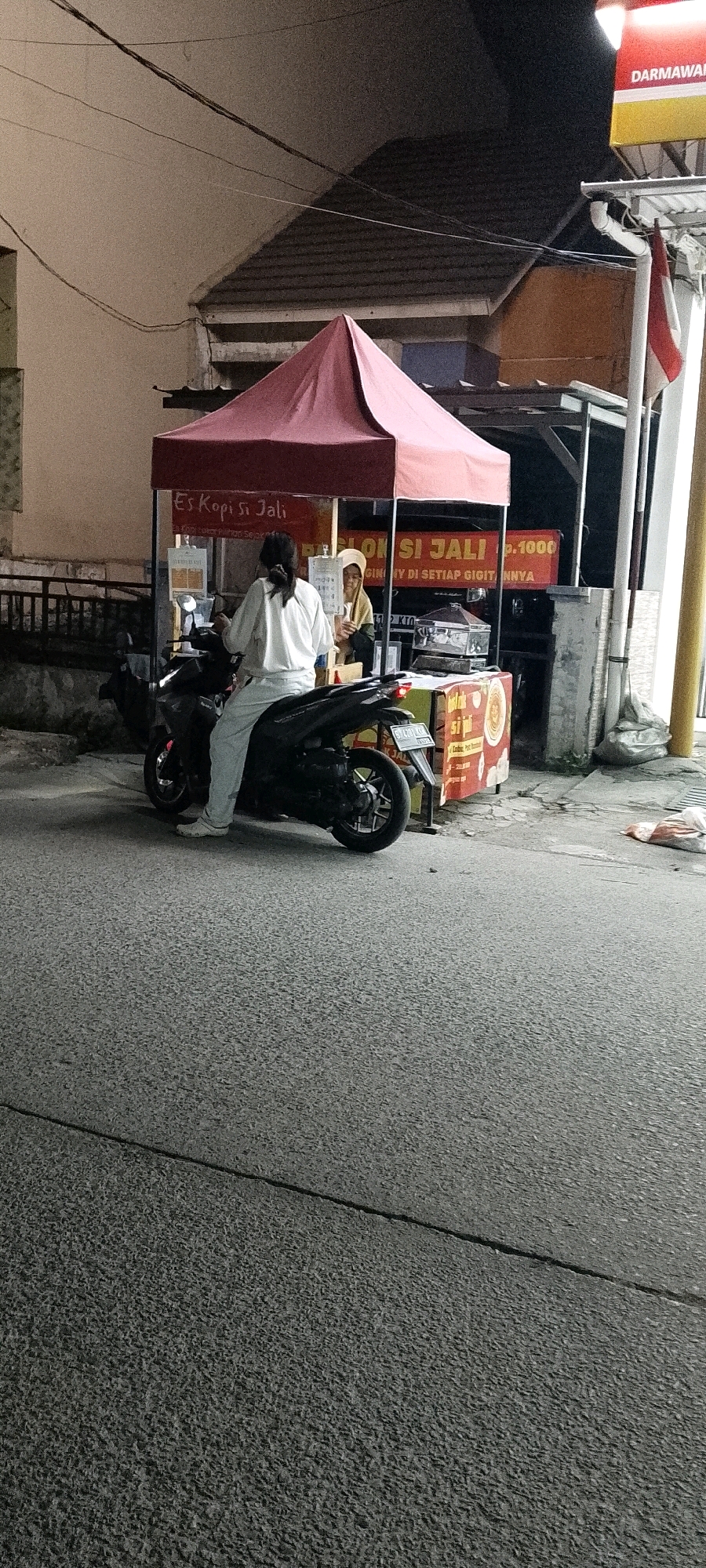 The image captures a night scene in Satriamekar, Indonesia, where a person is seated on a black motorcycle in front of a food stall. The stall has a red awning and displays signage that reads "Es Kopi Si Jali" and "BABI JI JALI Rp.1000". Another individual, wearing a light-colored head covering, is visible behind the stall, possibly attending to a customer or preparing food. The motorcycle is parked on an asphalt road. The environment is dimly lit, suggesting it is nighttime, with lights from the stall and a nearby building illuminating the area. The building in the background appears to have a tiled roof. A flag, possibly the Indonesian flag, is hanging from a pole on the right side of the image. Various plastic bags and debris are scattered on the ground near the stall.