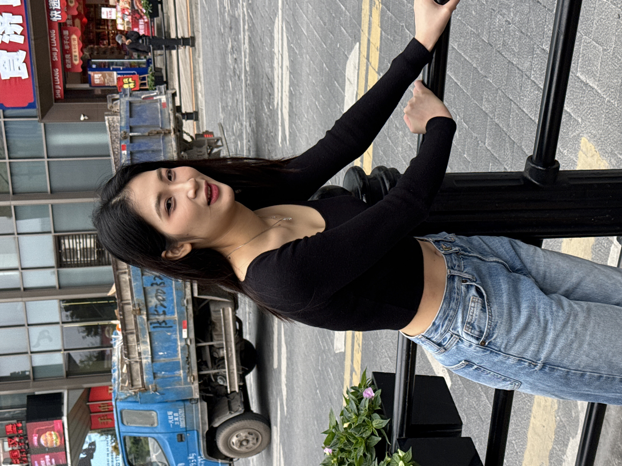 Here is a description of the image:
A woman is posing for a photograph in the Yuzhong District of China. She has long, dark hair and is wearing a long-sleeved black top and jeans. She is holding onto a black metal railing.
Behind her, there is a street with a truck and a building with Chinese characters on it. There are also some flower bushes.