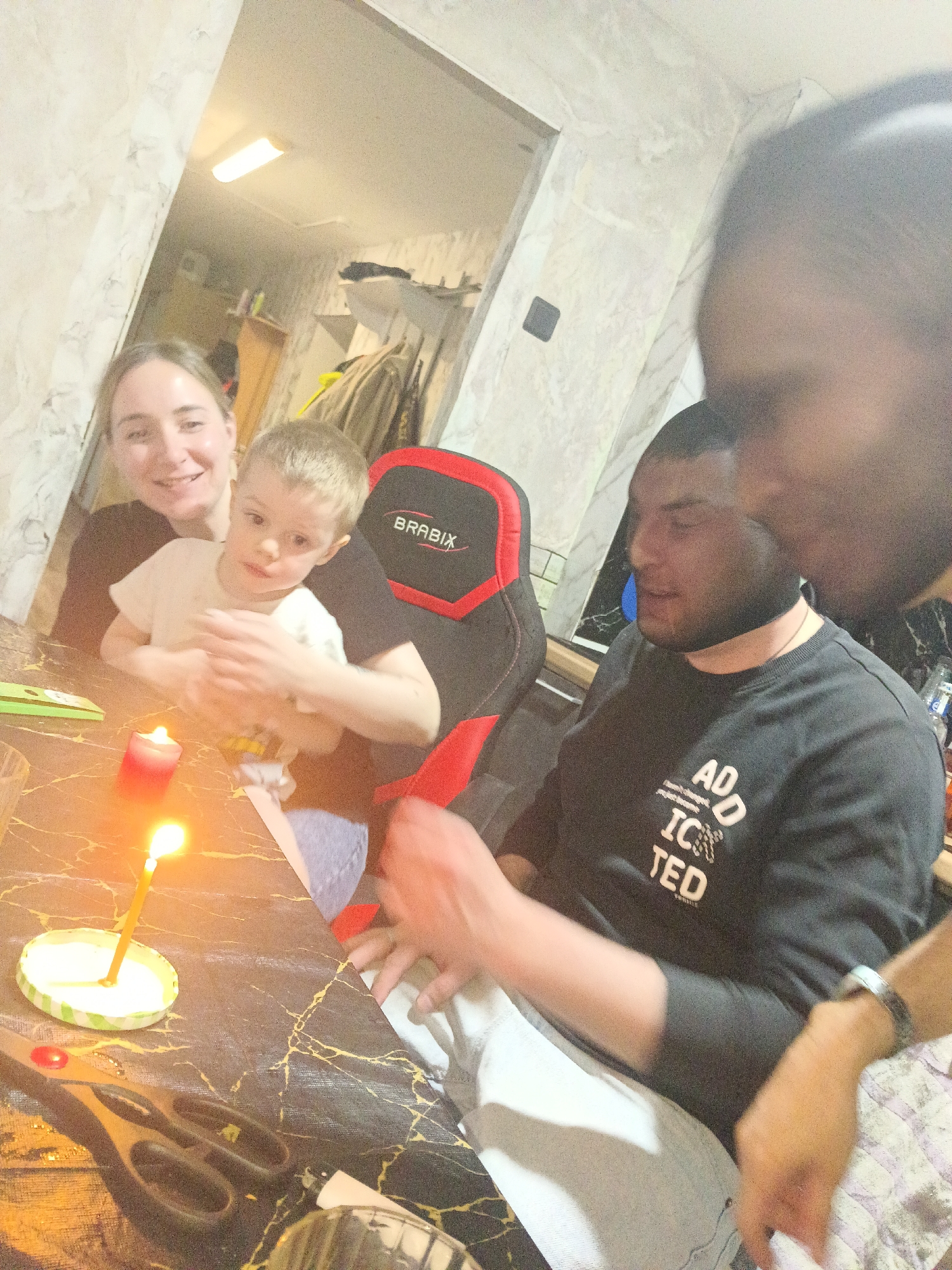 A medium shot captures a family gathered around a table. A woman and a young child sit near a black and red gaming chair labeled "BRABIX." A man wearing a black long-sleeved shirt with a white graphic sits at the table. Another individual stands very close to the camera. A candle burns in a small, makeshift holder, set on a black marble-patterned table. Scissors and other objects are also on the table. The location is Chaikovsky, Russia.