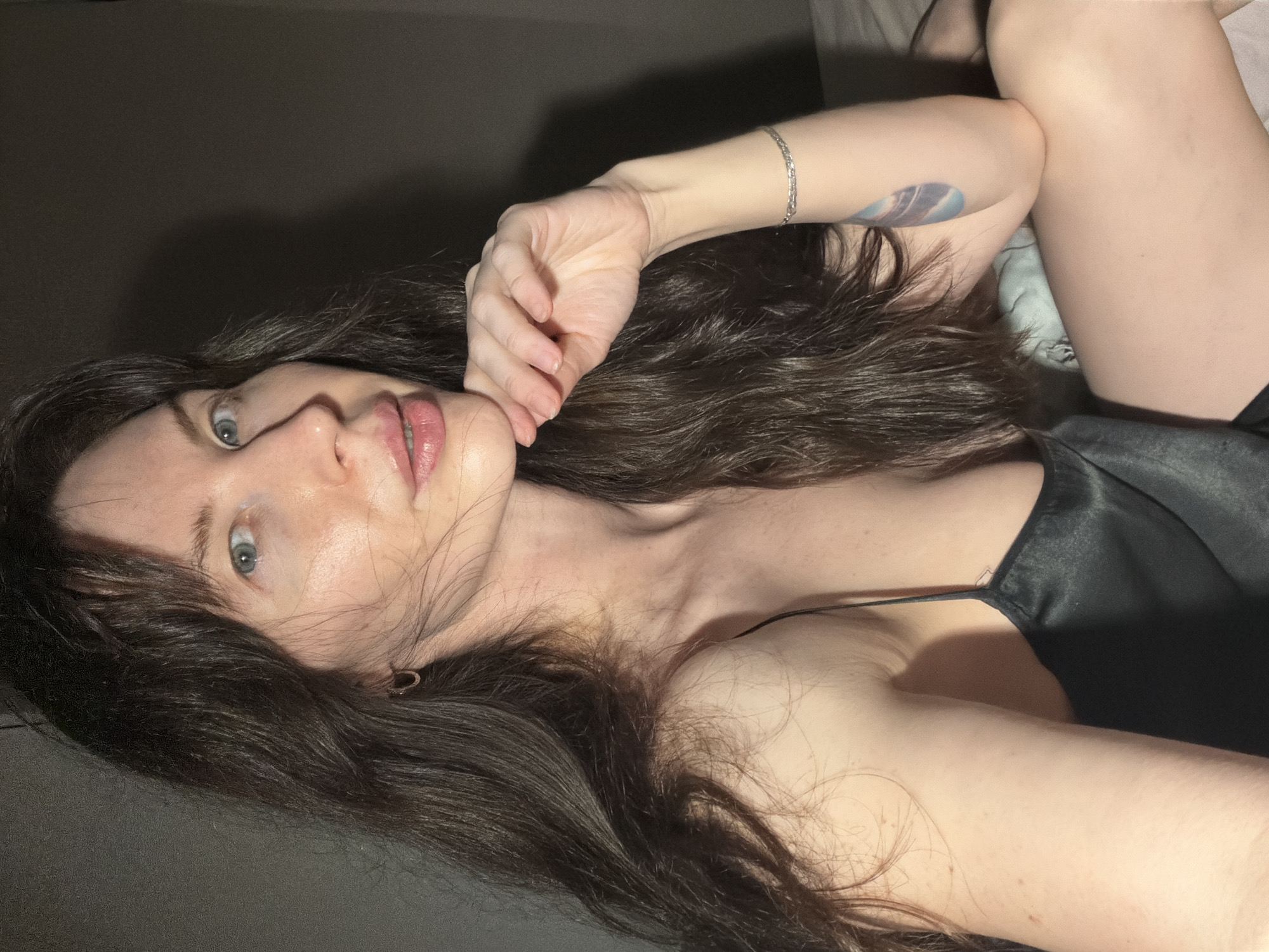 A woman with long, dark brown hair and blue eyes lies on what appears to be a dark gray or black surface, possibly a bed or sofa. She is wearing a dark-colored, possibly black, silk or satin camisole. Her left hand is raised near her face. A silver bracelet is visible on her left wrist, and there is a blue tattoo on her left forearm. The setting appears to be indoors, with subdued lighting and a dark background. There is no information about the specific location in Perm, Russia, or any actions, interactions, or environment details beyond what is immediately visible in the image.