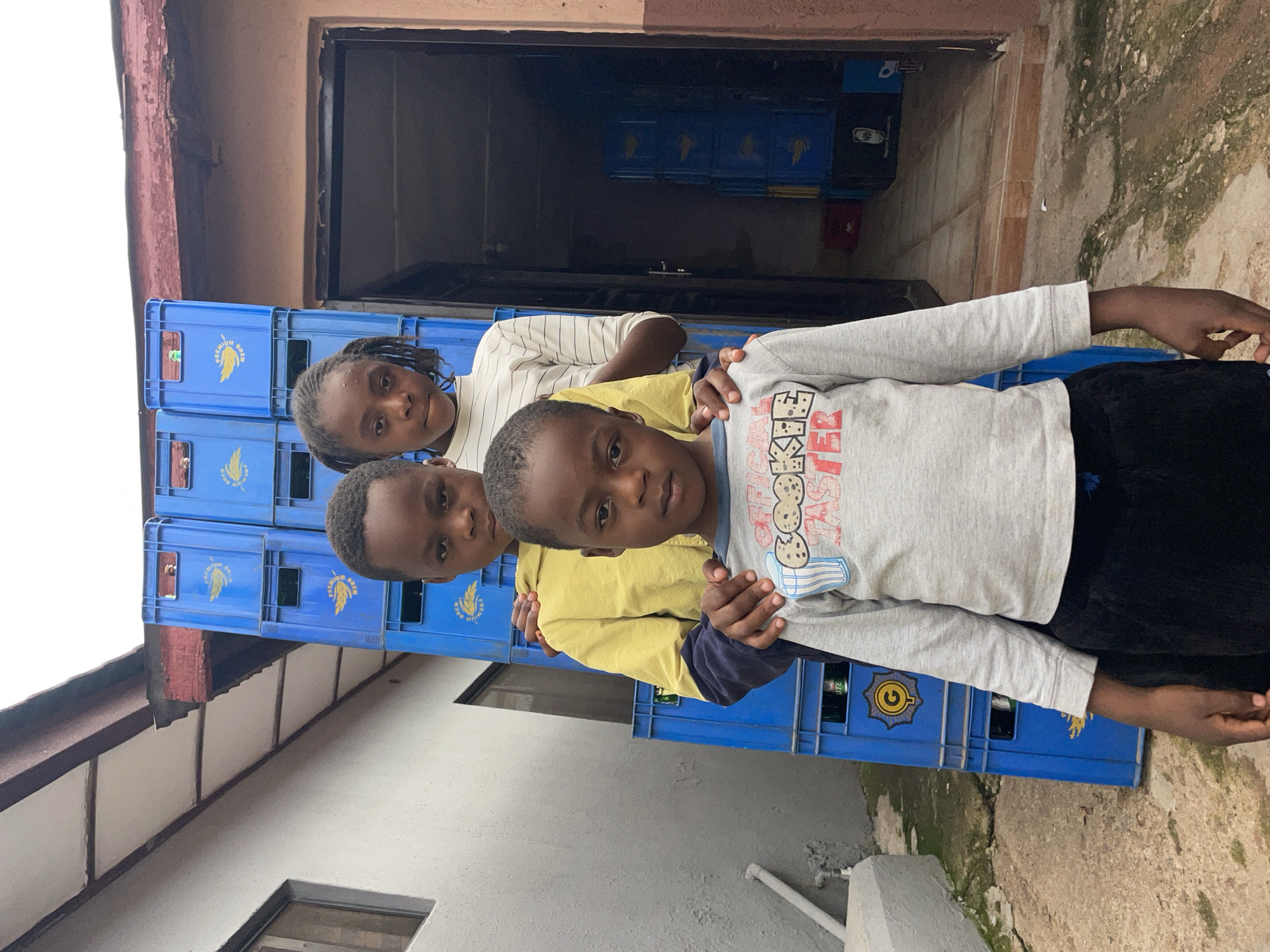 In Jos, Nigeria, three children stand outdoors in front of stacked blue crates. The foreground features a young boy, approximately 4-6 years old, facing forward with a neutral expression. He wears a grey long-sleeve shirt with red text, including "COOKIE MONSTER," and dark pants. Behind him, a second boy, approximately 6-8 years old, stands with his left hand on the first boy's right shoulder and his right hand on the shoulder of the girl behind him. This second boy wears a yellow and dark blue long-sleeve top and also looks forward with a neutral expression. The third child, a girl approximately 8-10 years old with dark braided pigtails, stands behind the second boy, looking directly at the camera. She wears a white and grey striped long-sleeve shirt.
Behind the children, a tall stack of blue plastic crates dominates the background, extending from the left to the center-right. Each crate displays a golden winged emblem and text such as "PREMIUM BREW." Some crates visibly contain bottles. To the far left, a light-colored building wall with a window is partially visible, featuring a white pipe running vertically. Above the crates and to the right, a weathered reddish-brown corrugated metal roof or awning is present. To the right of the main crate stack, a dark doorway leads into a building, revealing more blue crates stacked within the interior. The ground is an uneven surface of concrete or packed earth with patches of green moss or vegetation. The sky above is uniformly light grey, indicating an overcast condition.
