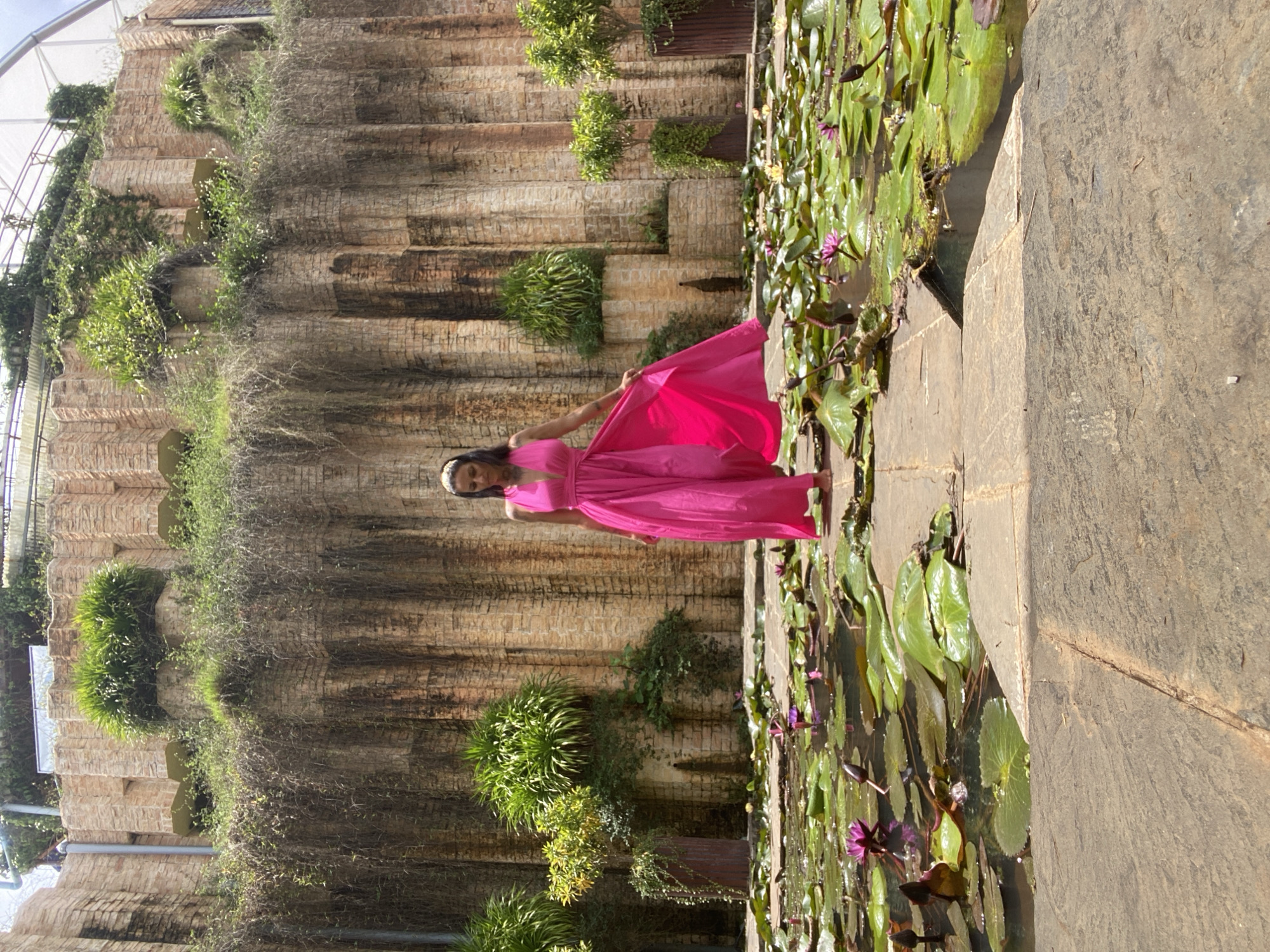 This image depicts a woman in a bright pink dress standing on a stone pathway in front of a pond filled with water lilies. The pond is bordered by stone, and behind it is a large, tiered structure made of light brown brickwork covered in lush greenery. The brickwork has a vertical, column-like texture. The woman is wearing a headband and her dress is long and flowing, with a deep V-neck. She appears to be walking or posing. The water lilies in the pond have green pads and some have pink blossoms. The background suggests an outdoor, possibly botanical garden or park setting, consistent with locations like Cam Ly in Da Lat, Vietnam, known for its natural beauty and gardens.