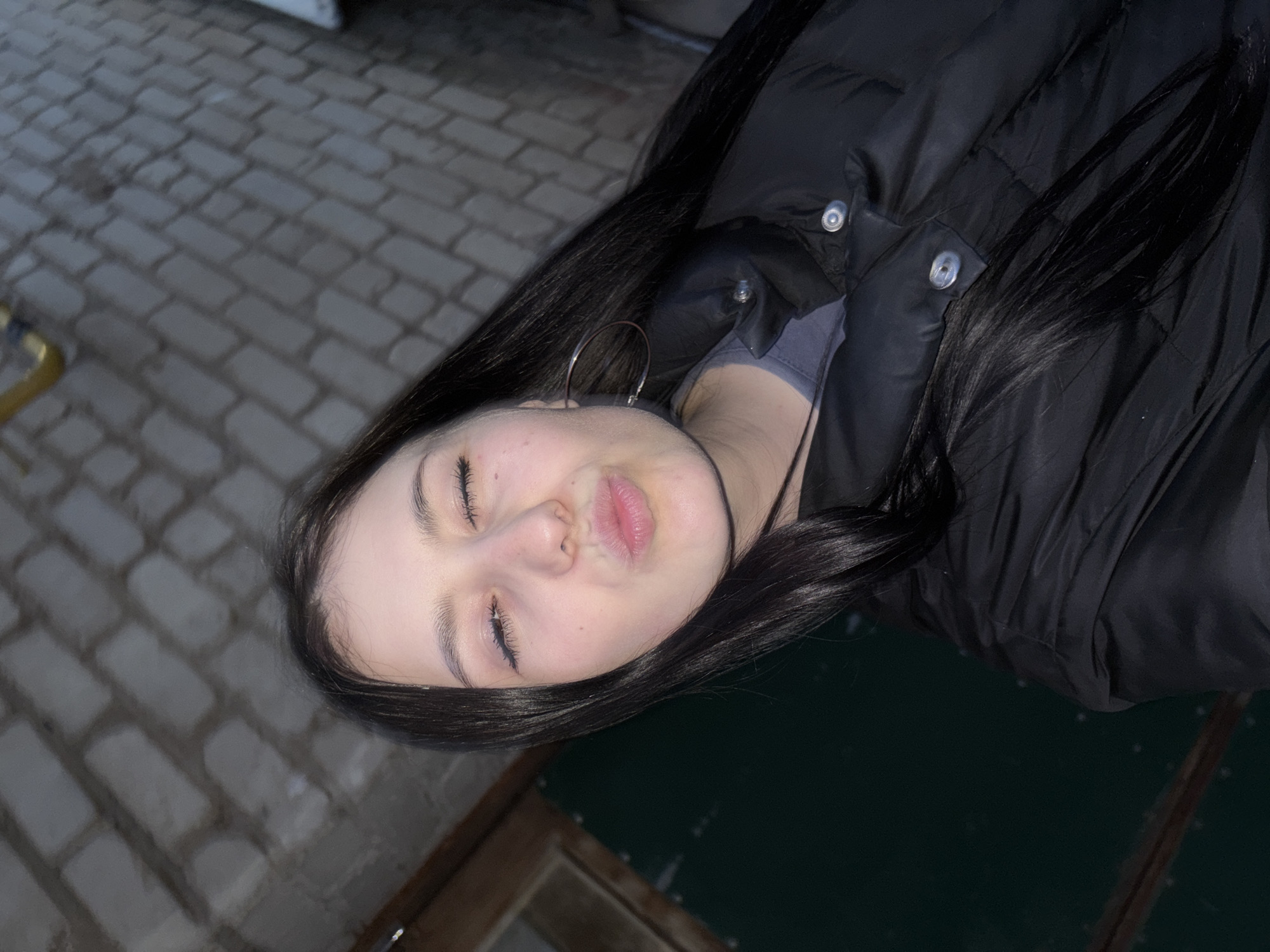 A young woman with long, dark hair, wearing a black puffer jacket and silver hoop earrings, is positioned in the foreground. Her eyes are closed, and her lips are slightly pursed. To the left of the woman, there is a brick-paved ground, while on the lower right side, there is a dark surface, possibly a table or a dark-colored solid material. The photo appears to be taken outdoors in an urban setting. The location is Фоки, Russia.