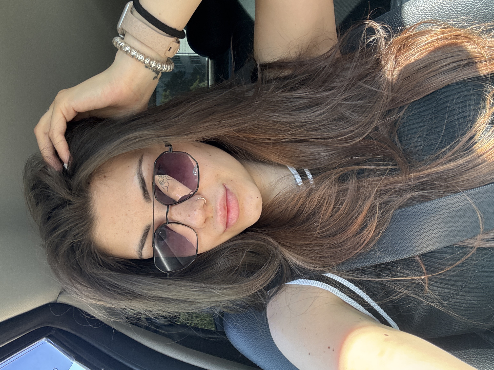 A young woman with long, dark brown hair and fair skin is sitting in the passenger seat of a car, looking at the camera. She is wearing sunglasses, a nose ring, and several bracelets on her left wrist. Her hair is partially covering her face. The car interior is visible, with a black leather seat and a seatbelt across her body. The background suggests an outdoor setting, possibly in the city of Sofia, Bulgaria, with glimpses of trees and buildings through the car window. The sunlight is casting shadows on her face and hair, indicating it is daytime.