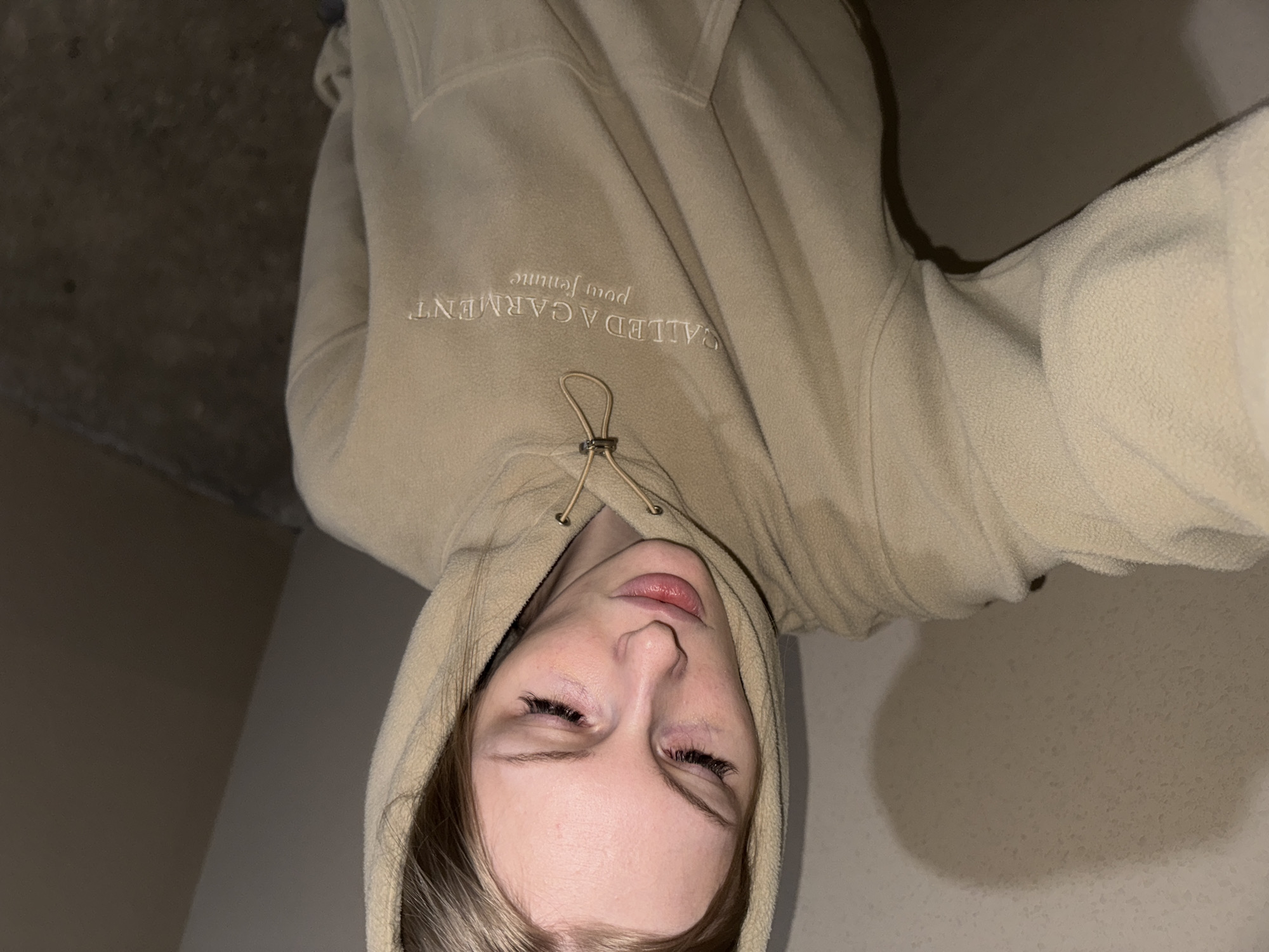 The image shows a close-up of a young woman wearing a beige hooded sweatshirt with "CALLED A GARMENT pour femme" embroidered on it. She has blonde hair and is looking down with her eyes closed. The lighting is dim, and the background appears to be a textured wall. There is no identifiable location in the city of Perm, Russia.