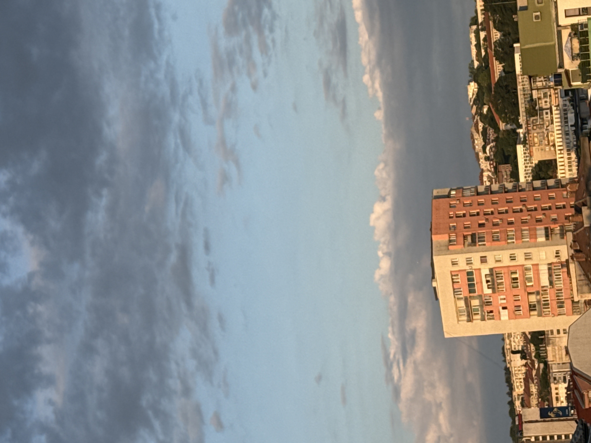 The image captures a cityscape with a prominent apartment building in the foreground, situated in Pleven, Bulgaria. The apartment building is tall and rectangular, painted in shades of orange and cream, with numerous windows arranged in a grid pattern. It is surrounded by other buildings of varying heights and architectural styles, some appearing older and more traditional with red-tiled roofs, while others are modern residential blocks. In the background, a hilly landscape dotted with trees and more buildings suggests the city extends into a more suburban or natural area. The sky above is a mix of blue and grey clouds, with some areas of soft, warm light, indicating it might be late afternoon or early evening. The overall scene is one of urban development in a hilly environment under a cloudy sky.