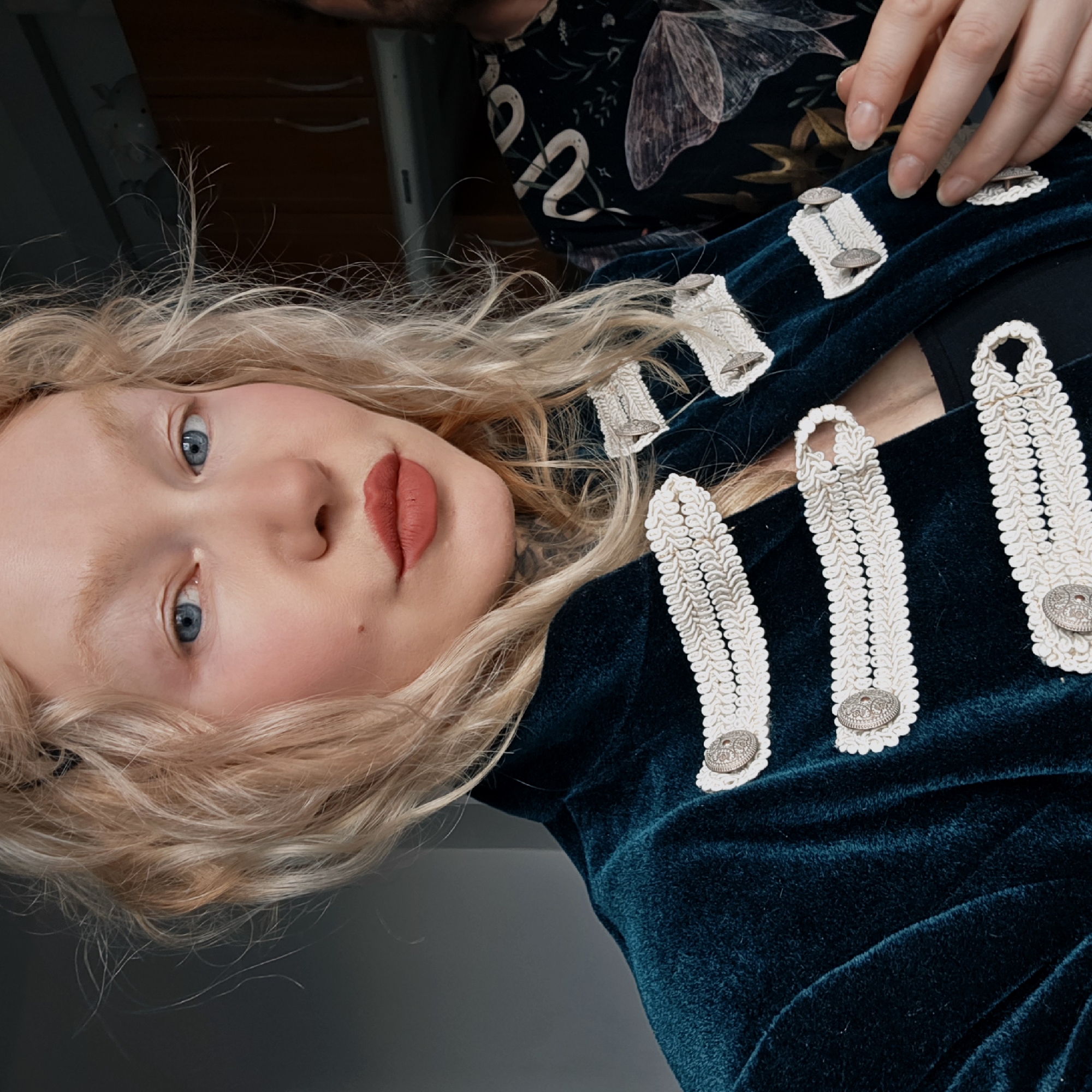The image shows a fair-skinned woman with blonde hair and blue eyes looking directly at the camera. She has light makeup, including red lipstick and blush. She is wearing a dark green velvet jacket with white braided details and silver buttons. A portion of her black bra is visible. In the background, there is a person wearing a dark, patterned shirt with what appears to be a snake and moth design. The setting is indoors. While it is not possible to confirm with certainty, nothing in the image specifically identifies the location as Saint Petersburg, Russia.