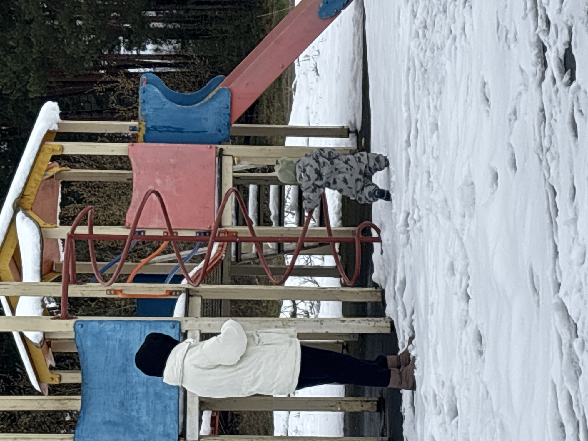 This image captures a winter scene at a snow-covered playground, likely in Razdolie, Russia. The ground is entirely covered in a thick, textured layer of white snow.
In the foreground, a multi-colored playground structure dominates the left side of the frame. It features wooden support beams, red metal rungs forming a ladder-like element, and panels painted in vibrant blue and red. A red slide descends from the upper platform of the structure. Snow accumulates on some of the horizontal surfaces of the playground equipment, contrasting with its bright colors.
Two individuals are visible. On a lower platform of the playground structure, an adult stands, viewed from behind. They are dressed in a white puffy jacket, black pants, a black beanie hat, and brown boots. Their posture suggests they are observing the activity in front of them.
To the right of the playground structure, a small child is positioned on the snow-covered ground. The child wears a grey and white camouflage-patterned snowsuit, a light green or grey hat, and dark footwear. They are in a crouching or leaning posture, seemingly playing in the snow or interacting with the base of the playground equipment.
The background consists of a dense forest of bare trees, typical of a winter landscape. The lighting indicates it is daytime, likely overcast, contributing to the diffused light. No text is visible in the image. The scene conveys a moment of outdoor activity during a cold, snowy day.