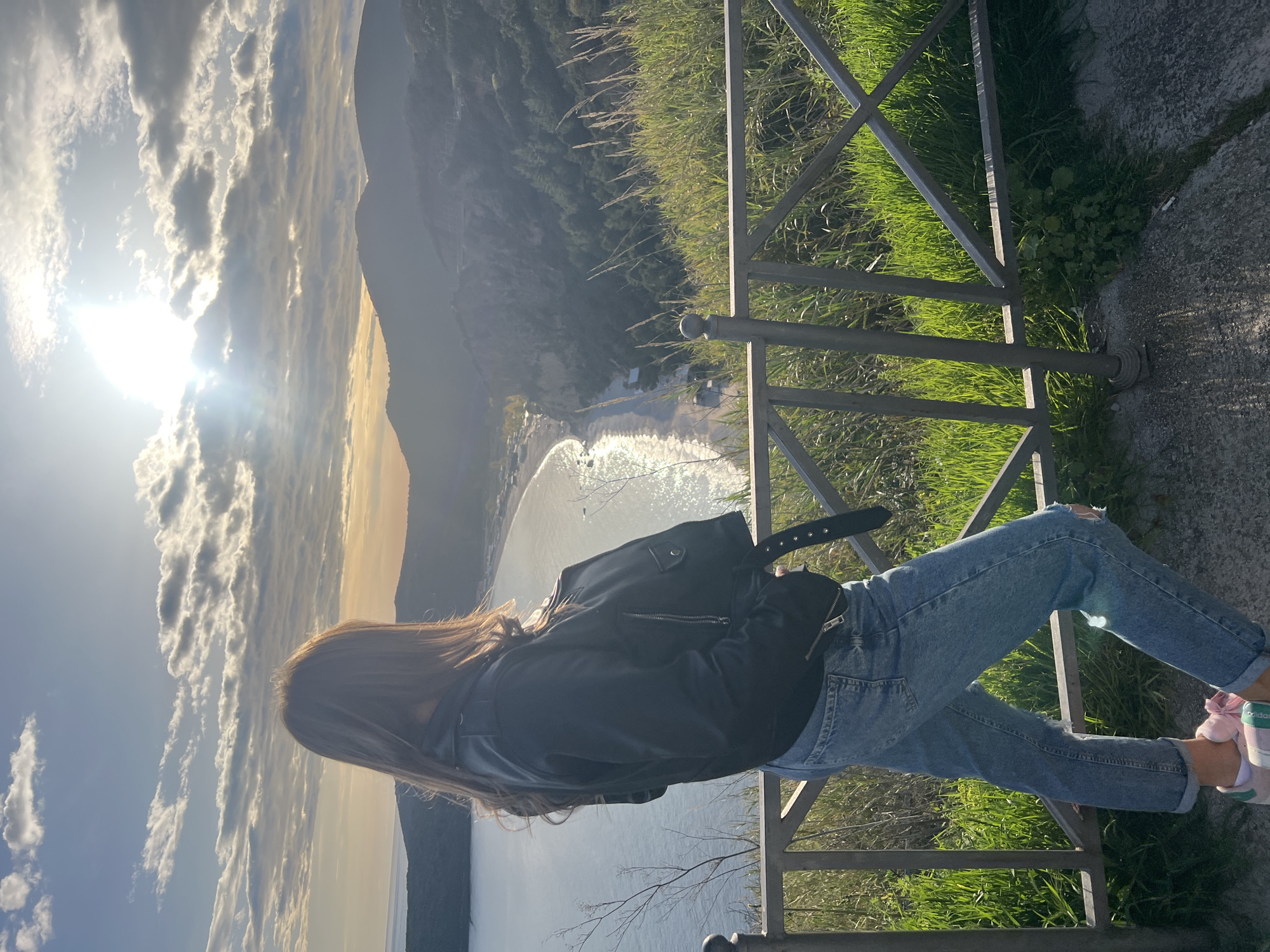 A full body shot of a woman is positioned on a stone overlook in Prijevor, Montenegro. She is wearing a black jacket and blue jeans, and is standing next to a silver metal rail fence with green grass and foliage behind it. Her back is to the camera. In the distance, a body of water and sun glare can be seen. Mountains are visible in the far background. The sky is covered in clouds.