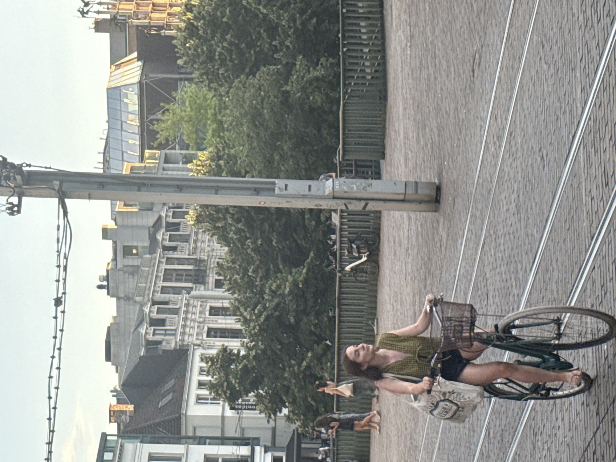 A woman rides a bicycle on a cobblestone street in Ghent, Belgium. The woman is wearing a green sleeveless top and black shorts, with a canvas bag slung over her left shoulder. Her bicycle is dark green and has a basket on the front. In the background, there are several buildings with ornate architecture and trees. A lamppost with wires runs vertically in the foreground. The tram tracks are visible on the street. The scene appears to be during the late afternoon or early evening, given the lighting.