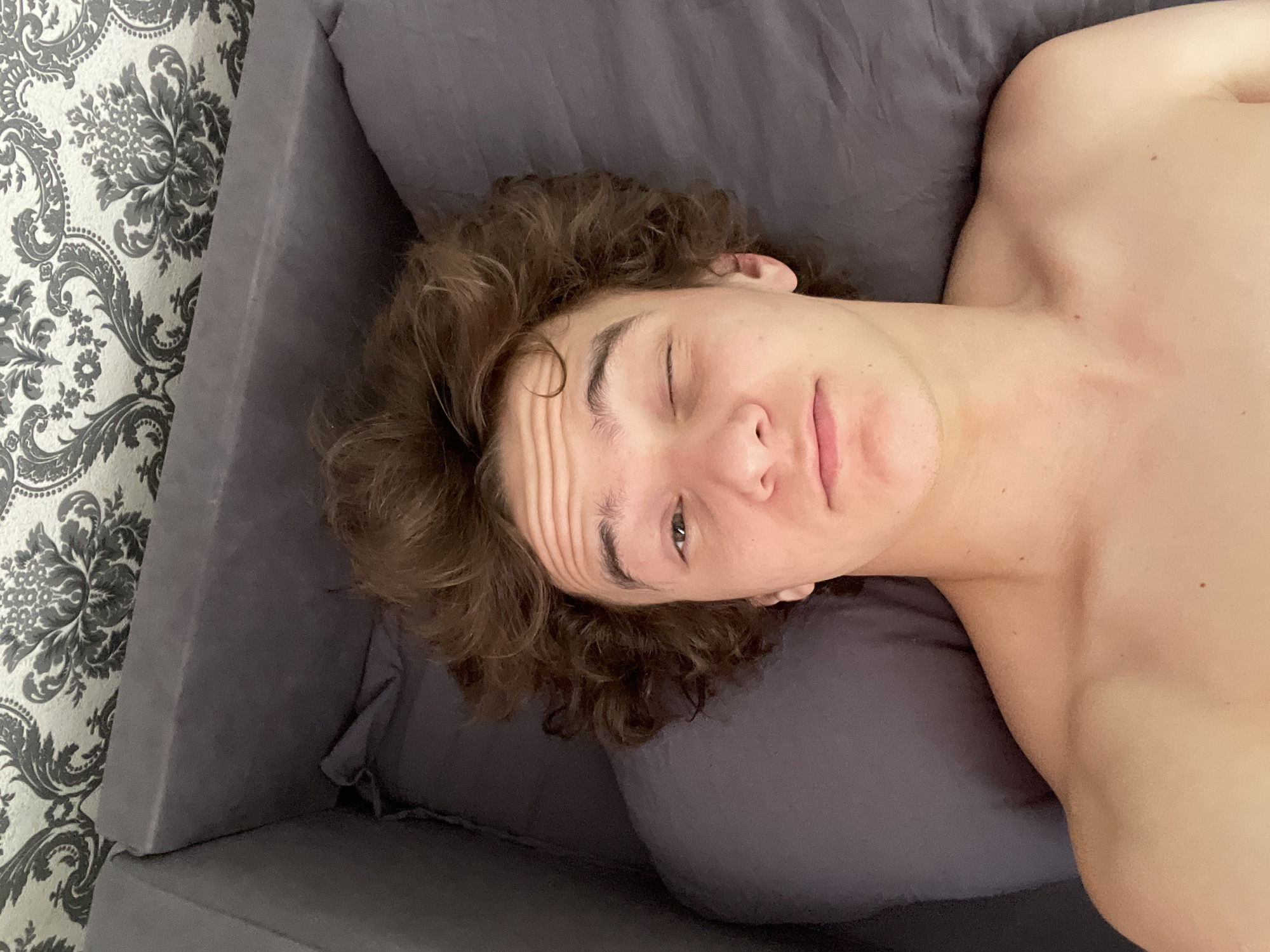 A shirtless young man with curly brown hair is lying on a gray couch. His head is on a gray pillow, and his left eye is closed. The background includes a wall with gray and white floral patterned wallpaper and the edge of the gray couch. There is no information to determine the location in Krasnoyarsk, Russia.