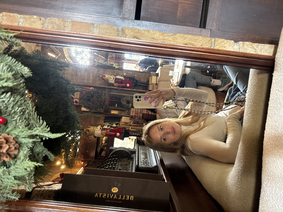A young adult female with light-colored hair, wearing a white turtleneck and blue jeans, takes a selfie in a large, framed mirror. She sits in a beige upholstered booth, holding a smartphone with a pink and white case and a patterned strap in her right hand, looking directly at the camera. The reflection reveals the interior of Caffetteria Belle Arti in Cagliari, Italy, which is decorated for the Christmas holiday season. Overhead, artificial fir branches with pinecones and red berries are visible. Within the reflected cafe, two large decorative nutcracker figures stand prominently, one primarily red and gold, and the other red, blue, and gold. Additional Christmas wreaths and wrapped gift boxes are visible. To the left of the female's reflection, a dark wooden display cabinet or counter holds a dark rectangular box with the inverted text "BELLE ARTI" visible. The cafe features exposed brick walls and wooden accents. Natural light enters through a window with a lattice pattern. In the deeper reflection, other patrons are visible, including one person wearing blue jeans and shoes with a visible Nike logo, seated at a small round table.