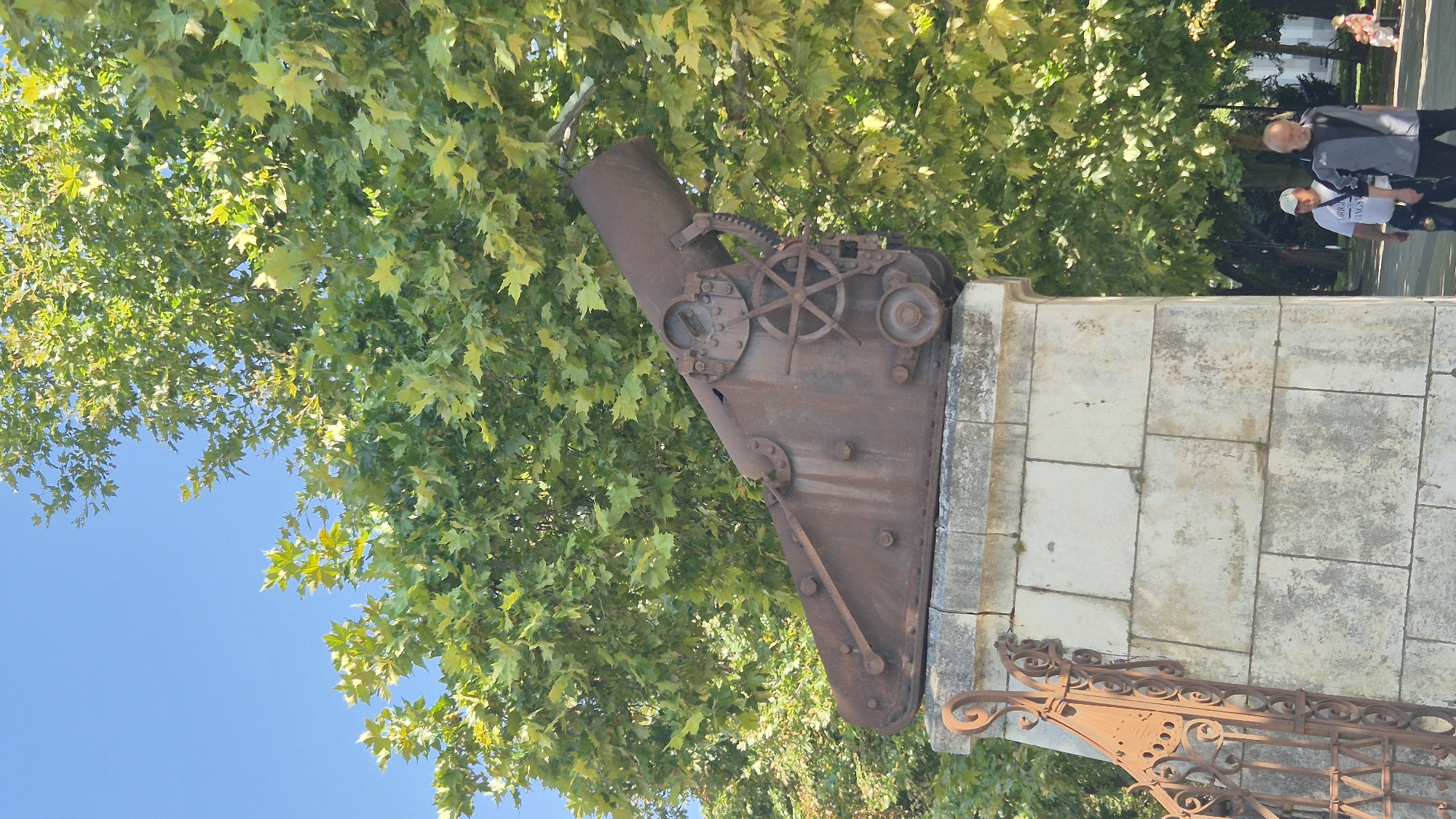 A large, rusted cannon is mounted on a stone pedestal in a public park. The cannon, made of dark, weathered metal, appears to be an old artillery piece, possibly a mortar, with visible gears and mounting hardware. It is positioned outdoors, with lush green trees providing a leafy backdrop and a clear blue sky above. To the left of the pedestal, a decorative wrought-iron fence, also exhibiting rust, is visible. In the background, two men are walking, dressed casually in t-shirts and jackets. Further back, another person is seated on a bench. The environment suggests a well-maintained park area in a city, likely during daytime. This is a monument in the city of Pleven, Bulgaria, commemorating military history.