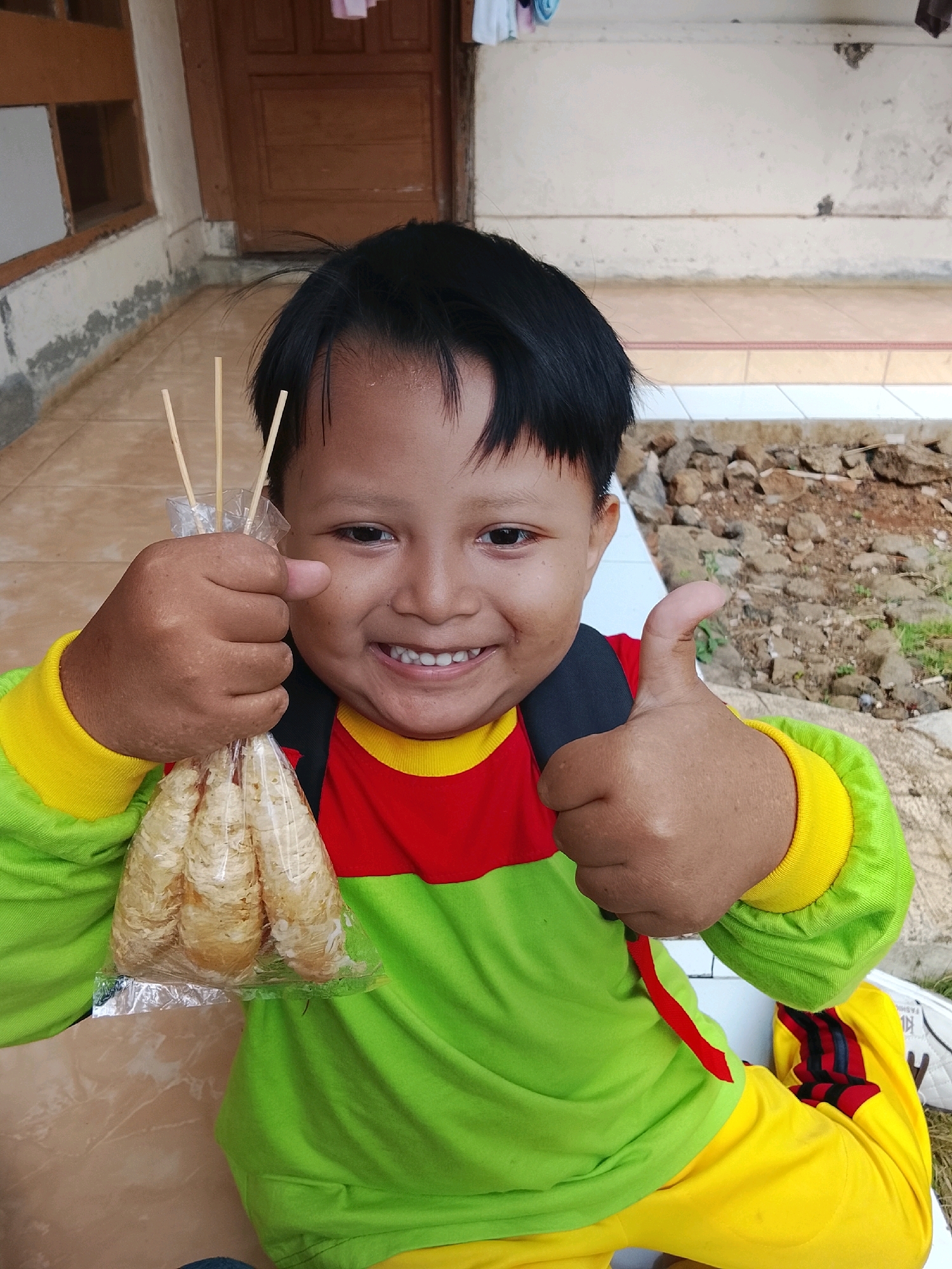 A young child, likely of Indonesian descent and appearing to be of primary school age, is captured in a medium close-up outdoor shot. The child is wearing a long-sleeved shirt with a vibrant green body, red chest panel, and yellow cuffs, paired with bright yellow pants featuring red and dark blue stripes. They are holding a clear plastic bag containing several food items, possibly snacks or sweets, on wooden skewers. The child's expression is cheerful, with a wide, toothy smile, and they are giving a thumbs-up gesture with their right hand. Their left hand is holding the bag of food. The background suggests a residential area, possibly in Kertahayu, Indonesia. To the left, a section of a building with wooden doors and windows is visible, along with a textured wall. The ground is a mix of paved areas and a rougher surface with small stones and dry leaves, indicative of a garden or yard. The lighting suggests it is daytime.