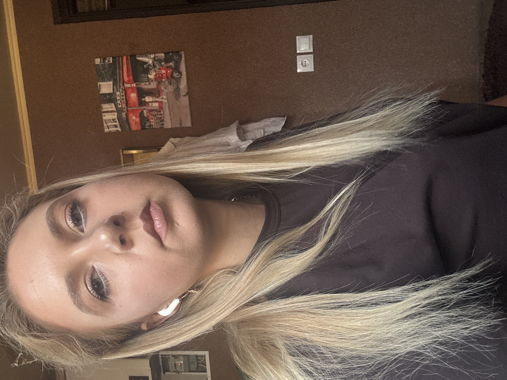 A young woman with blonde hair is the subject of this close-up, vertically oriented photograph. She is wearing a dark top and white wireless earbuds. Her face is visible, and she has makeup on, including eyeshadow and lipstick. The background consists of a corkboard wall with a framed picture of what appears to be a red fire truck and some buildings, as well as a white electrical outlet and light switch. The lighting suggests it is daytime, possibly indoors. The woman's expression is neutral.