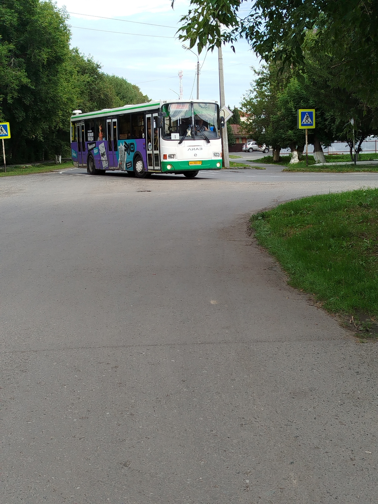 The image captures a public bus, a pedestrian crossing sign, and trees along a paved road in Zavodoukovsk, Russia. The bus is predominantly white with green and purple accents, featuring advertisements on its side. It appears to be in motion, driving on the road. A yellow pedestrian crossing sign with a black silhouette of a person is visible on the left side of the frame. The road is a mix of asphalt and possibly some lighter-colored paving. Trees with green foliage line the background, suggesting an urban or suburban environment. The sky is overcast with some visible clouds. The overall scene depicts a typical street view in a city.