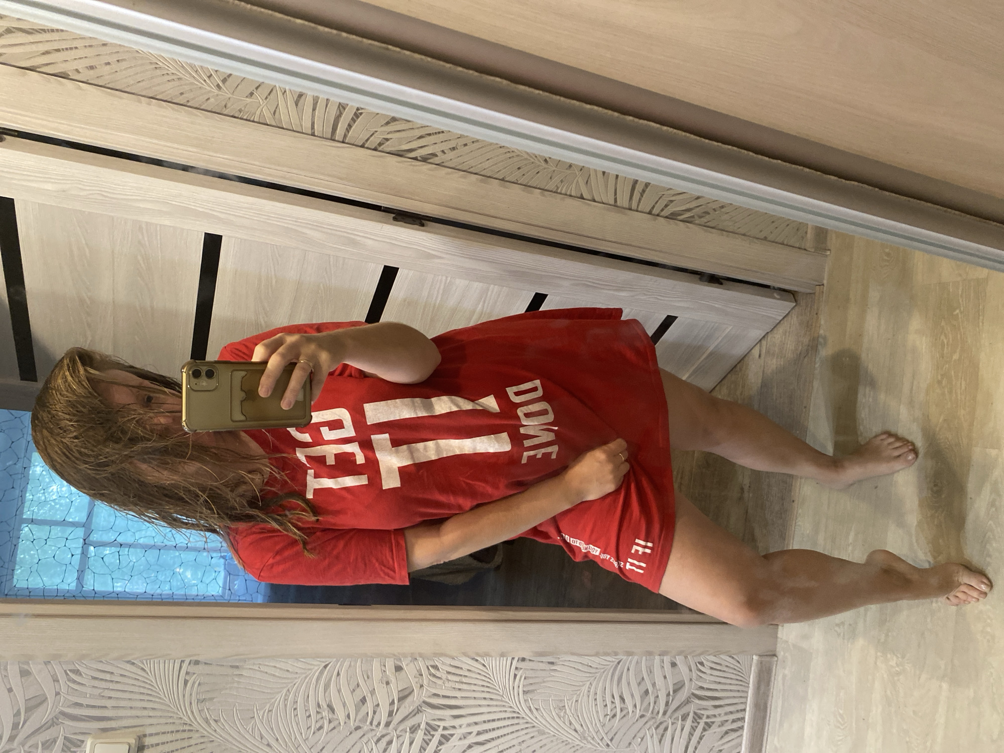A young woman with wet, sandy-blonde hair is taking a mirror selfie in a room. She is wearing an oversized red t-shirt with white text that appears to say "GET IT DONE" on the front. Her left hand is holding a gold-colored smartphone, and her right hand is lifting the hem of the t-shirt. She is barefoot and standing on a light-colored wood floor. The room has light wood doors and textured wallpaper with a palm leaf design. The overall impression is casual and intimate. It's impossible to determine the exact city location based on the image alone.